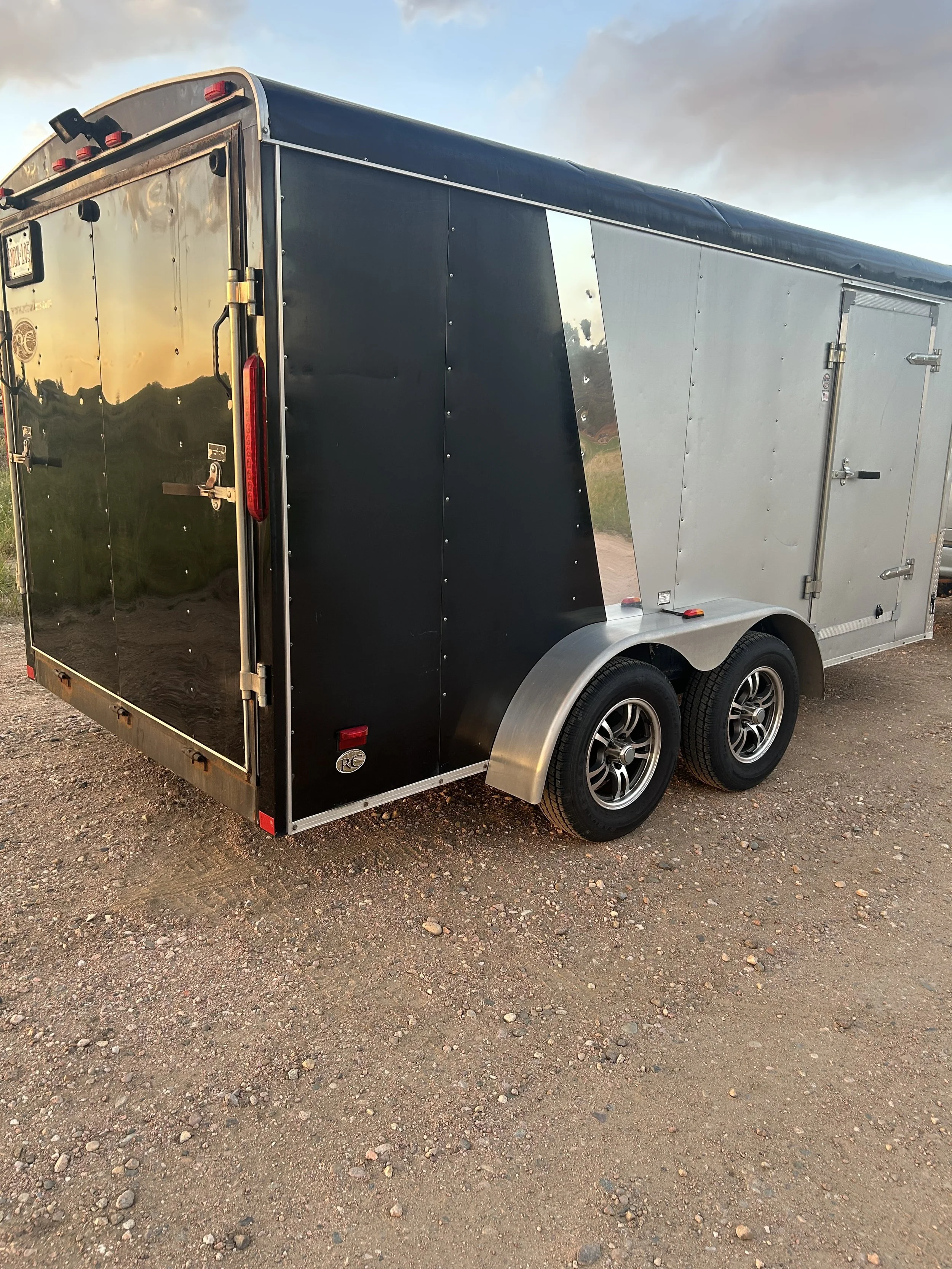 A double-axle enclosed cargo trailer with black and silver exterior parked on gravel ground.