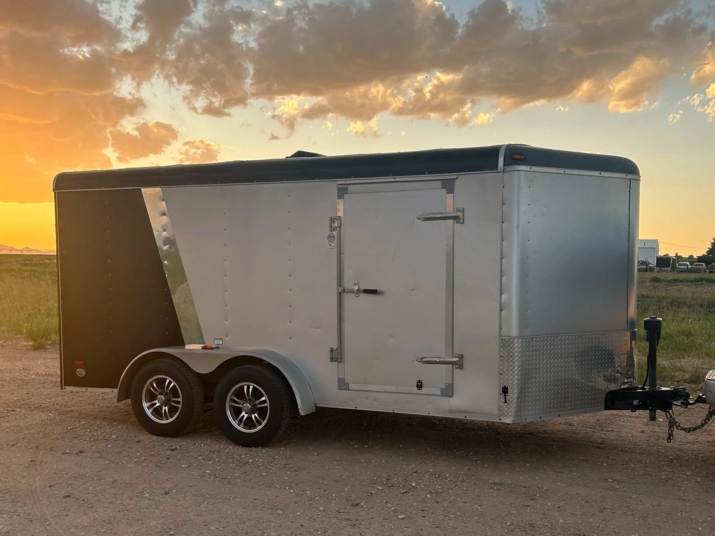 A secure enclosed enclosed trailer with two wheels parked on a dirt ground during sunset, with a sky filled with clouds and soft sunlight in the background.