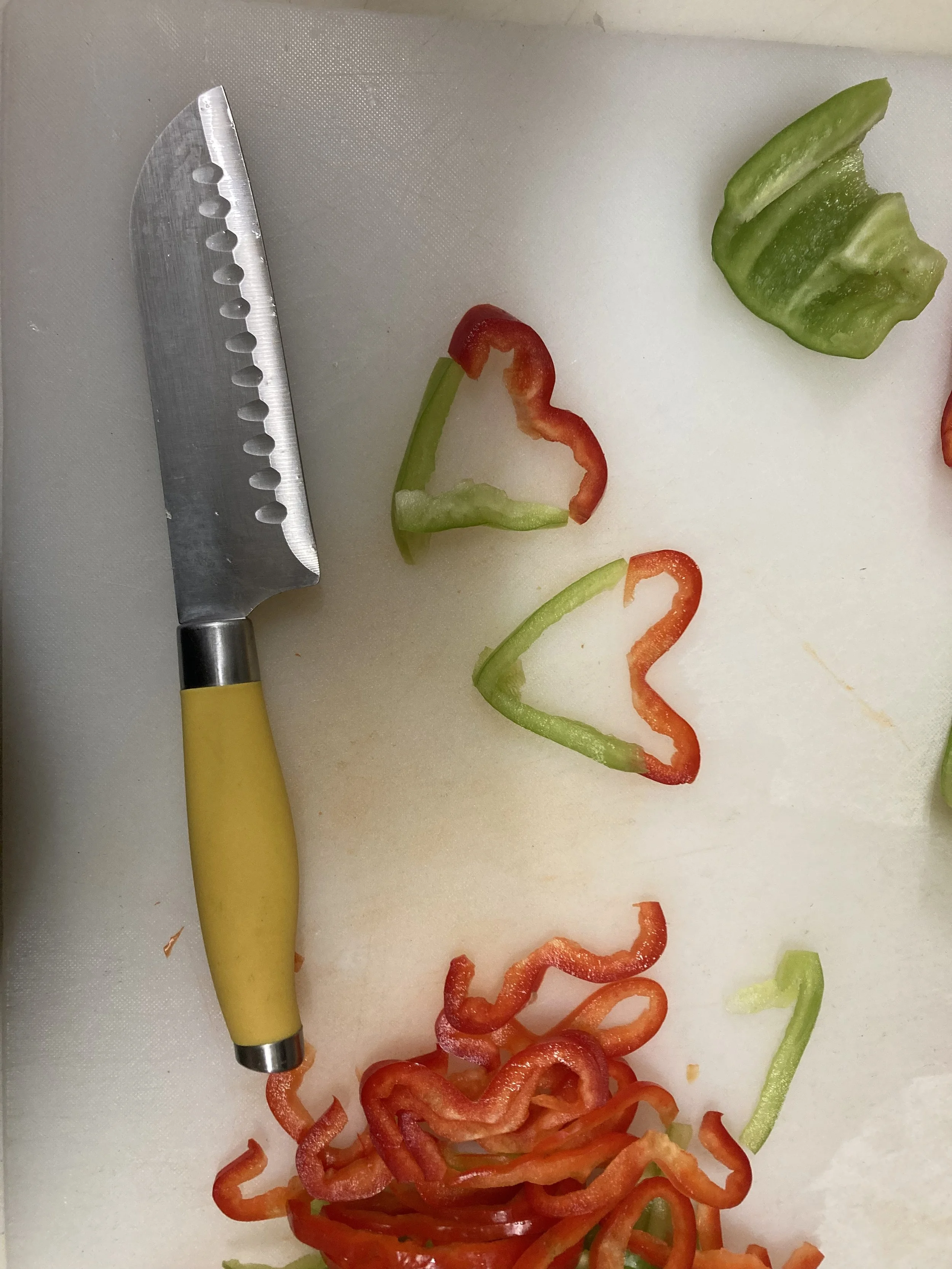 Sliced red, green, and orange bell peppers on a white cutting board with a yellow-handled chef's knife.