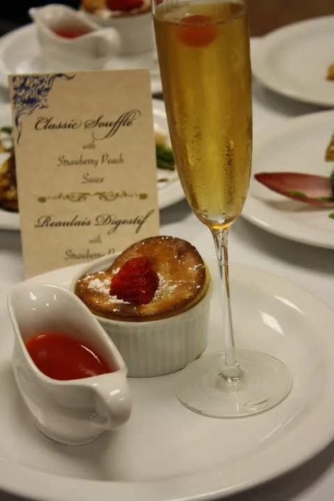A glass of champagne, a ramekin of strawberry sauce, and a baked dessert topped with strawberries on a white plate at a formal dining setting.