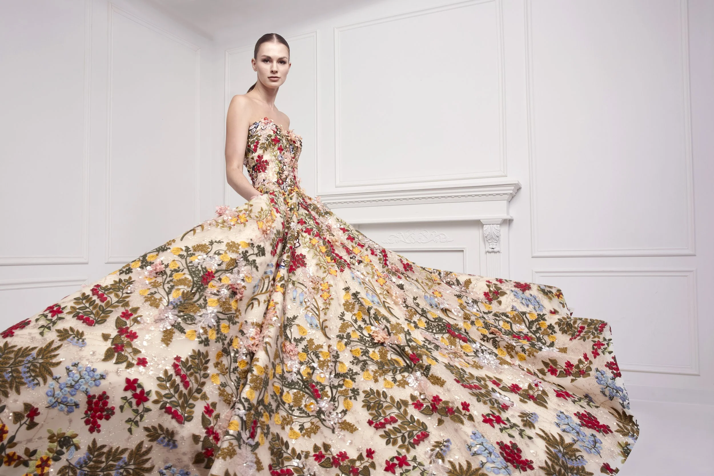 A woman wearing an elaborate, floral-embellished gown with a full train, standing in a white room with decorative wall paneling.