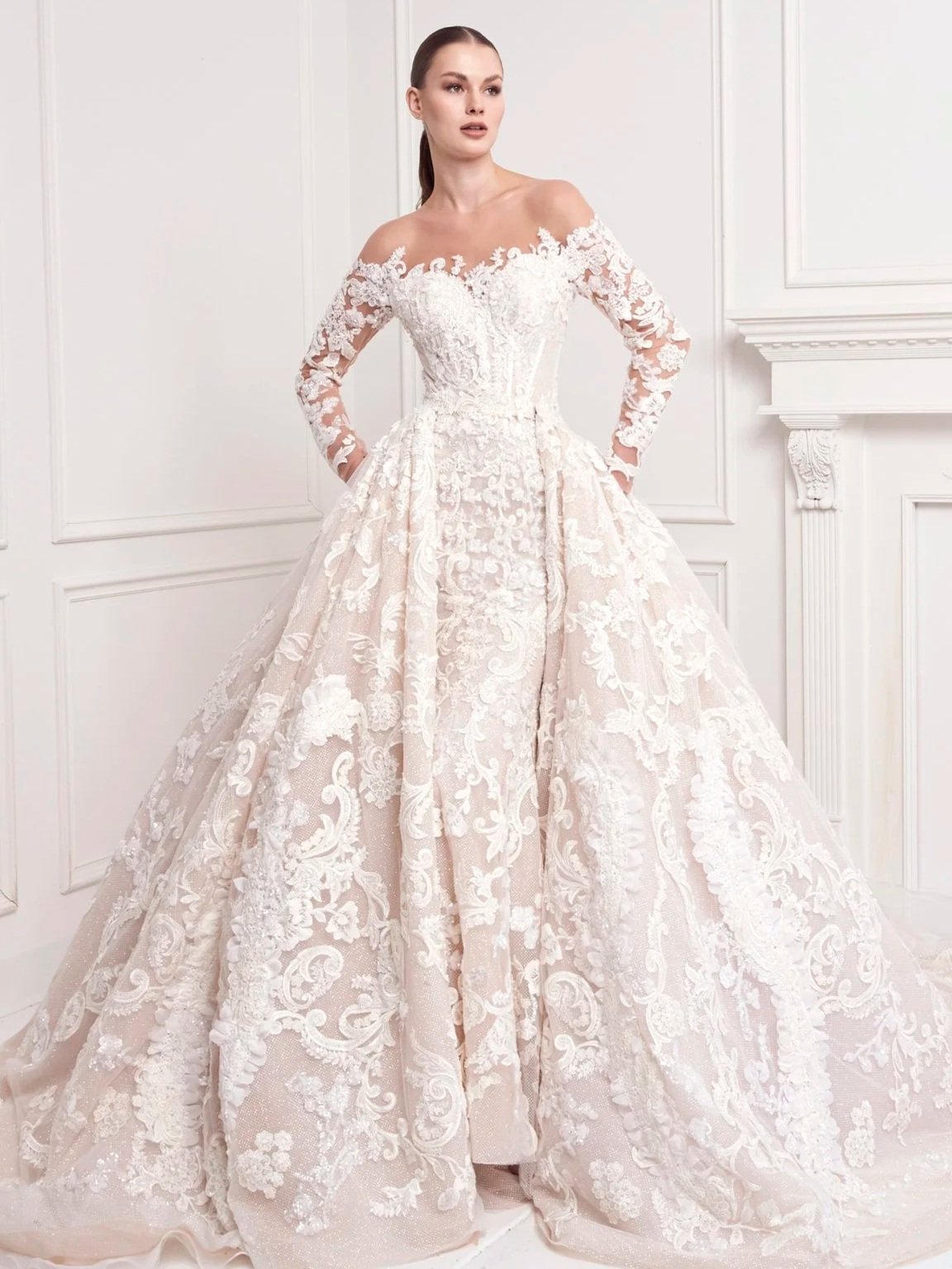A woman in an elaborate white wedding gown with lace embroidery, long sleeves, and a voluminous skirt, standing indoors against a white wall with decorative molding.