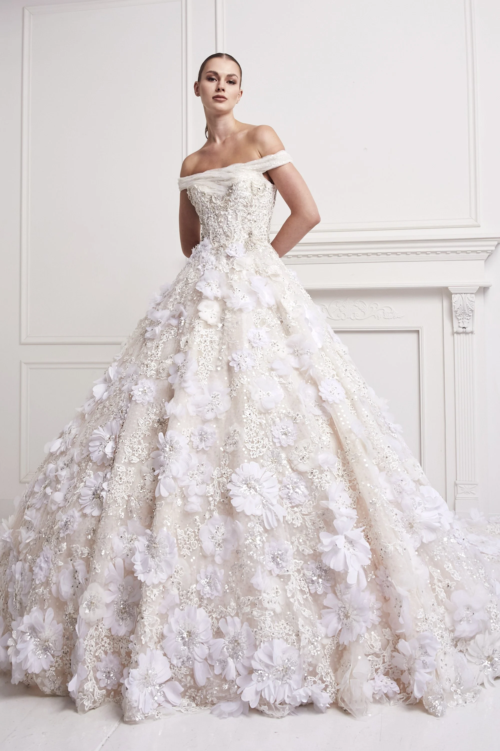 A woman in an off-the-shoulder white wedding gown with intricate lace and 3D floral appliqué details, standing in a room with white wainscoting and paneling.