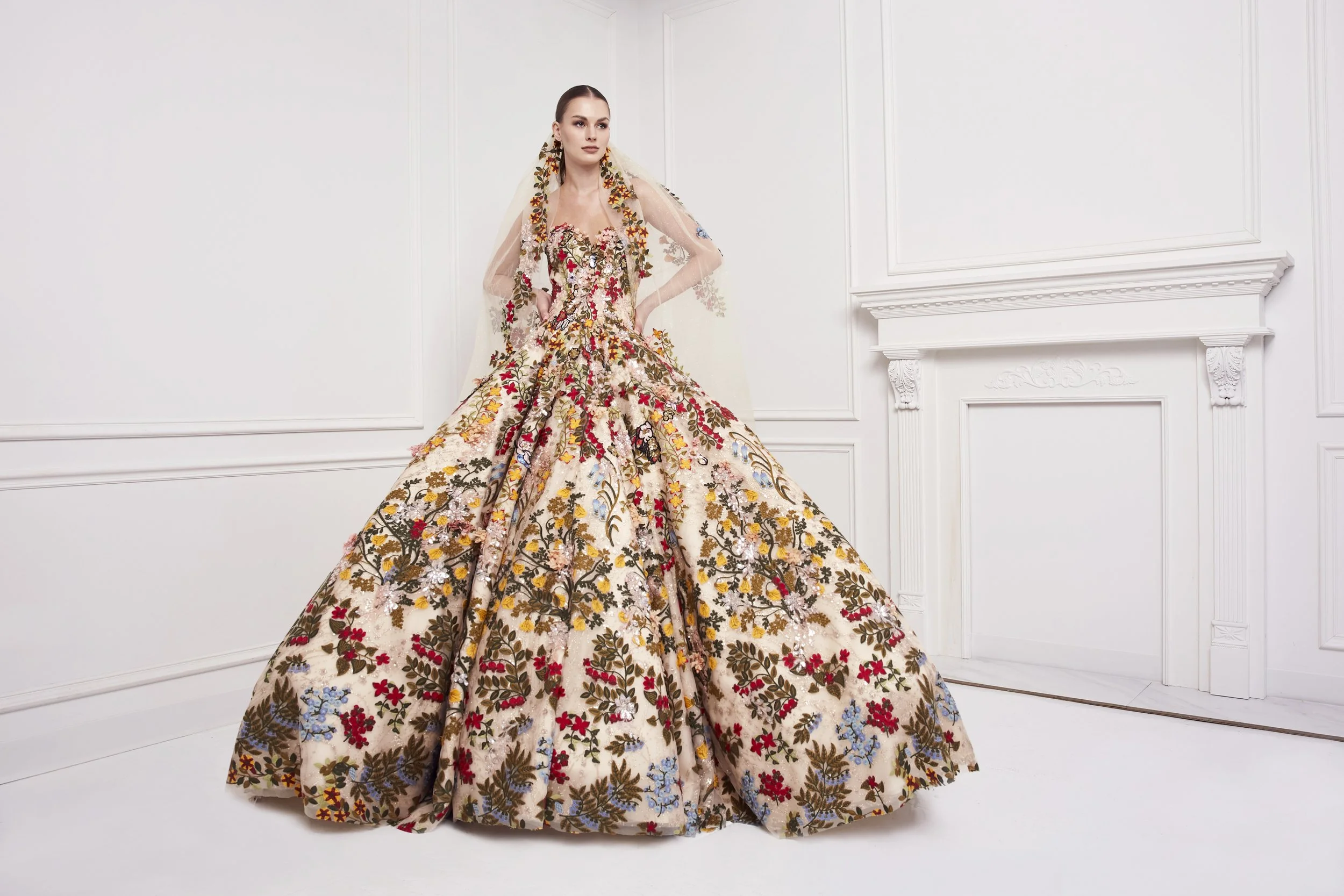 A woman standing in a white room wearing an ornate, long gown with intricate floral embroidery and a sheer cape with floral details.
