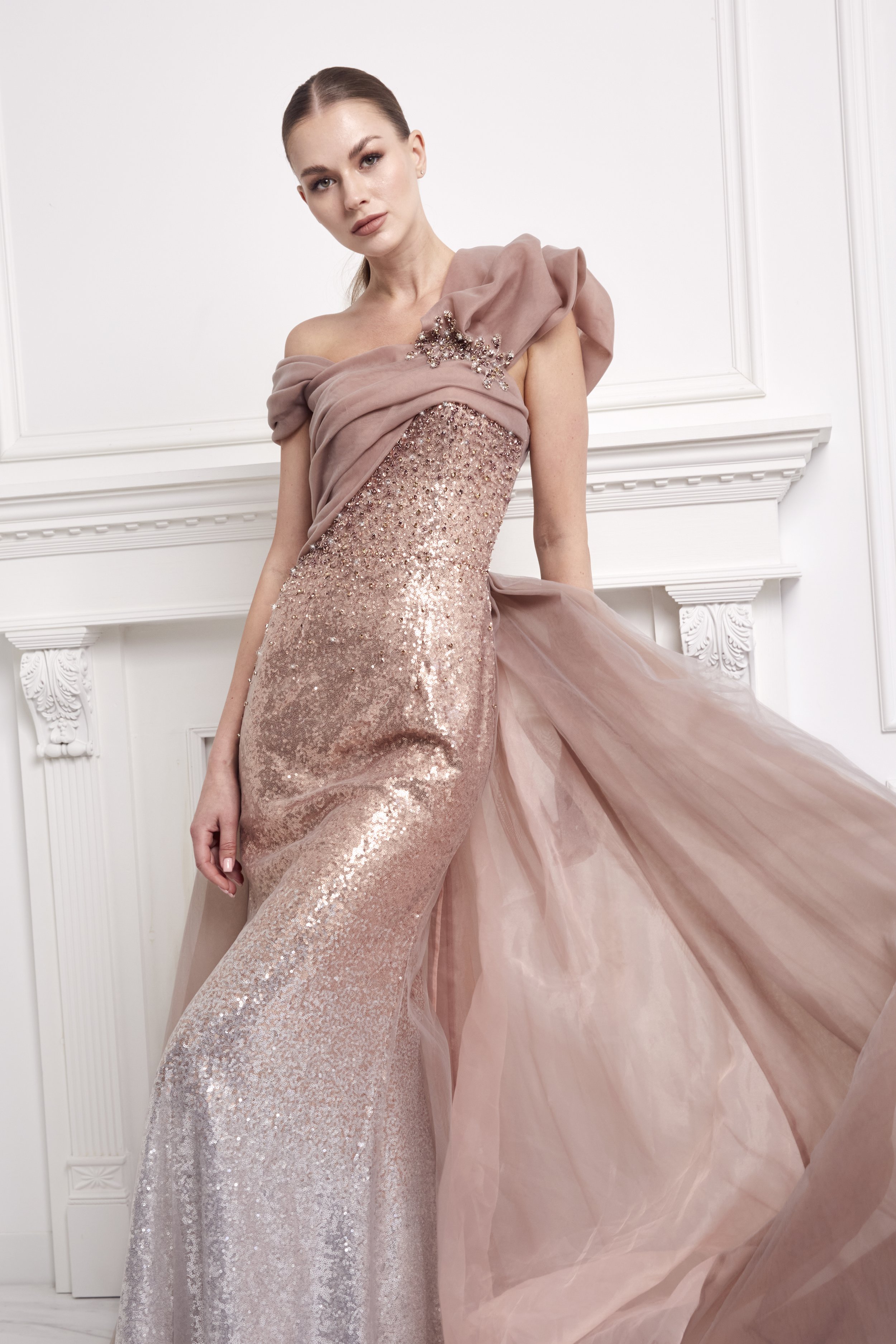 A woman in a strapless pink gown with sequin embellishments and a sheer draped overlay, standing in a white room with ornate molding.