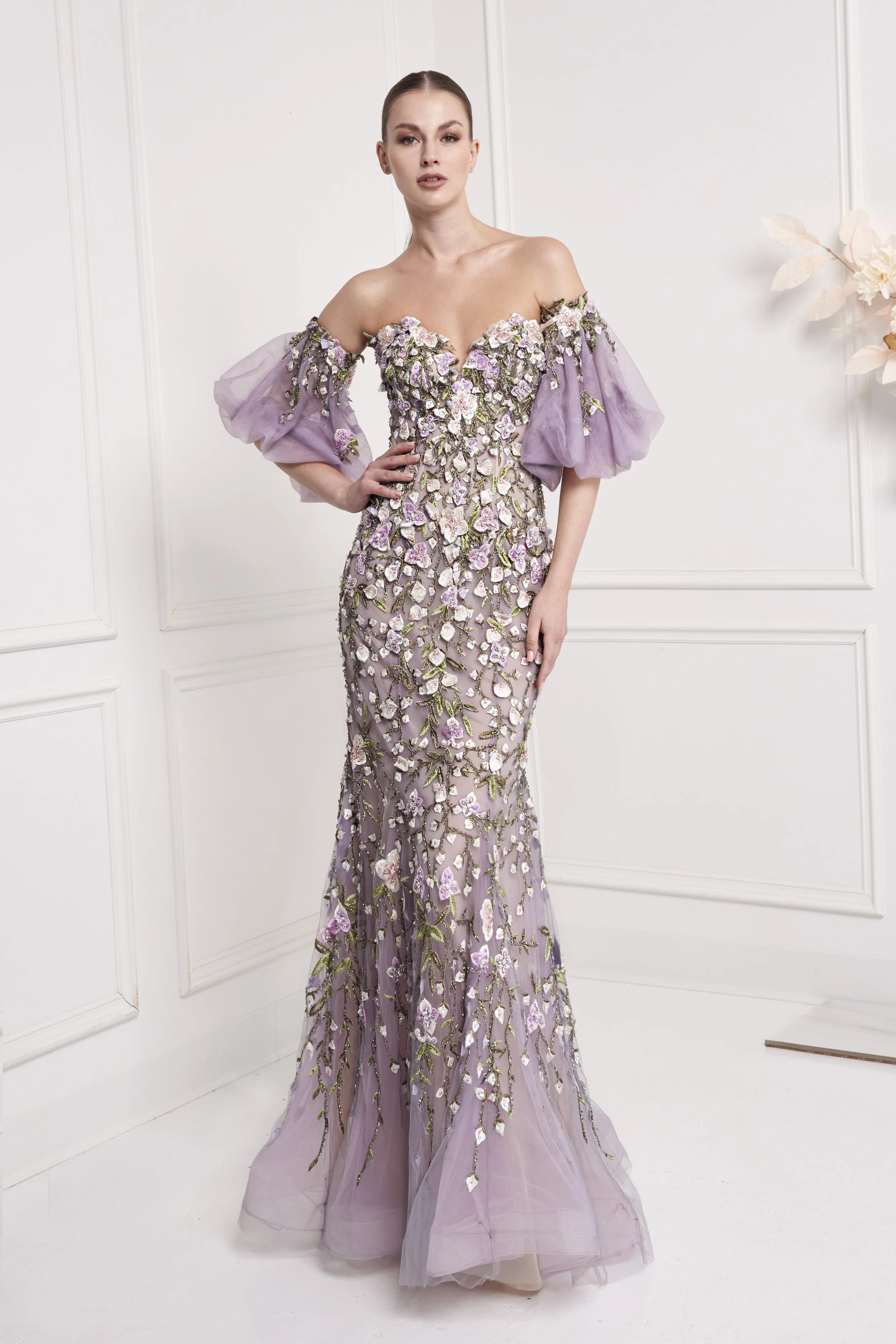 A woman wearing a lavender gown with floral embellishments, standing in a white room with panel molding, next to a flower arrangement.