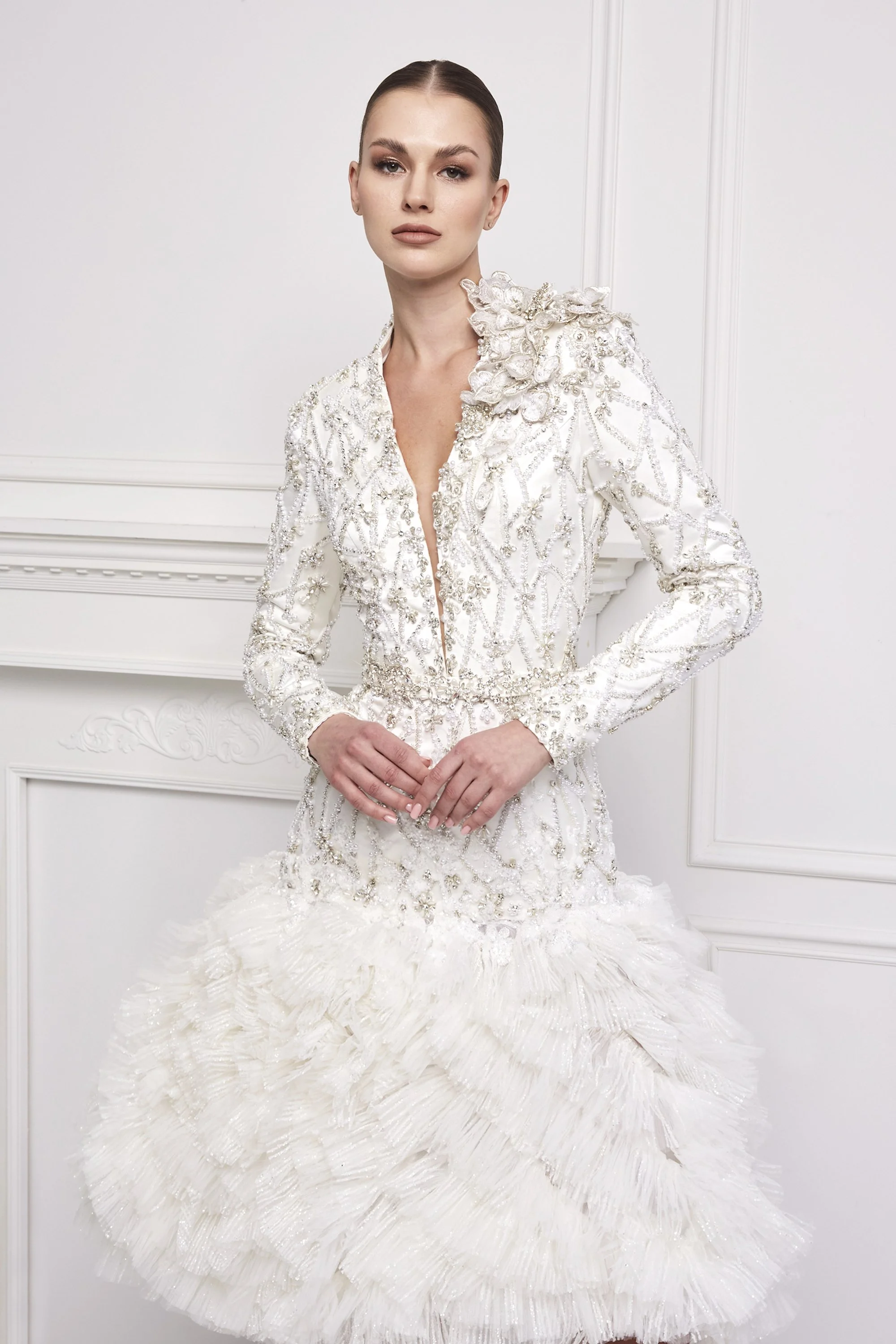 A woman in a white, ornate, embroidered wedding dress with floral appliqués and textured skirt, standing against a white paneled wall.