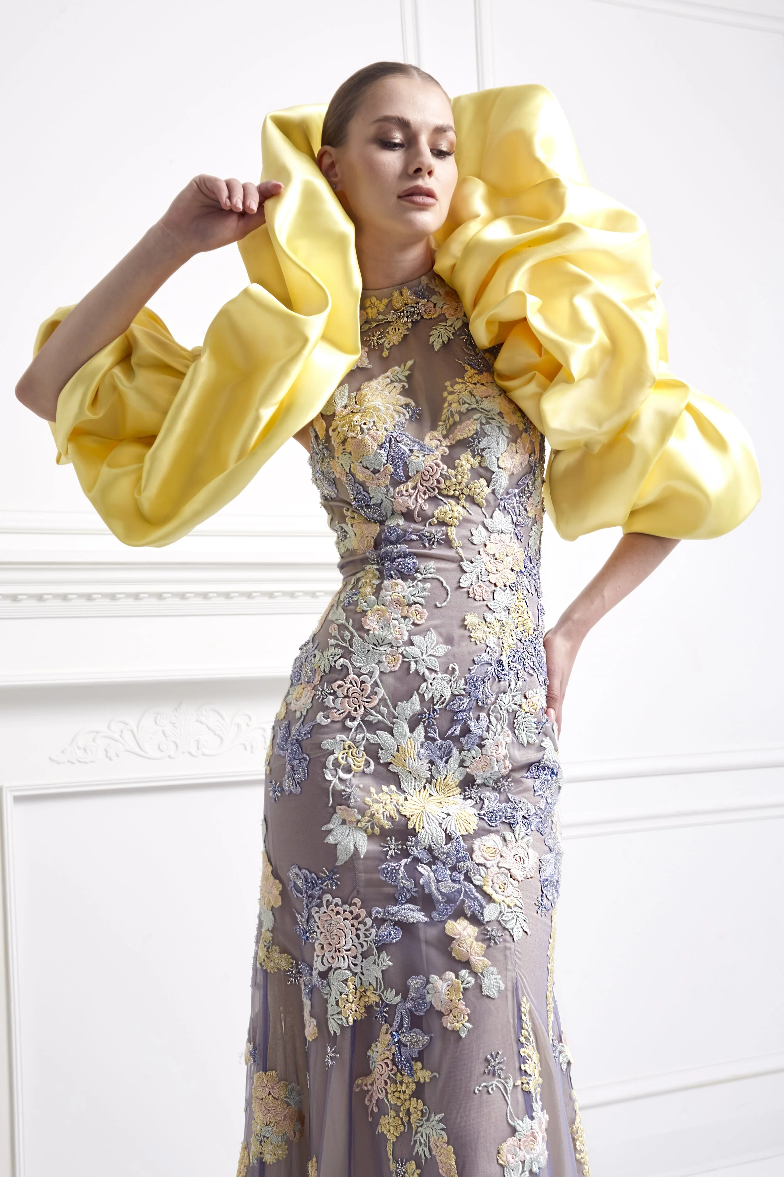 A woman in a floral embroidered evening gown with large yellow satin puffed sleeves, standing in a room with white walls and decorative molding.