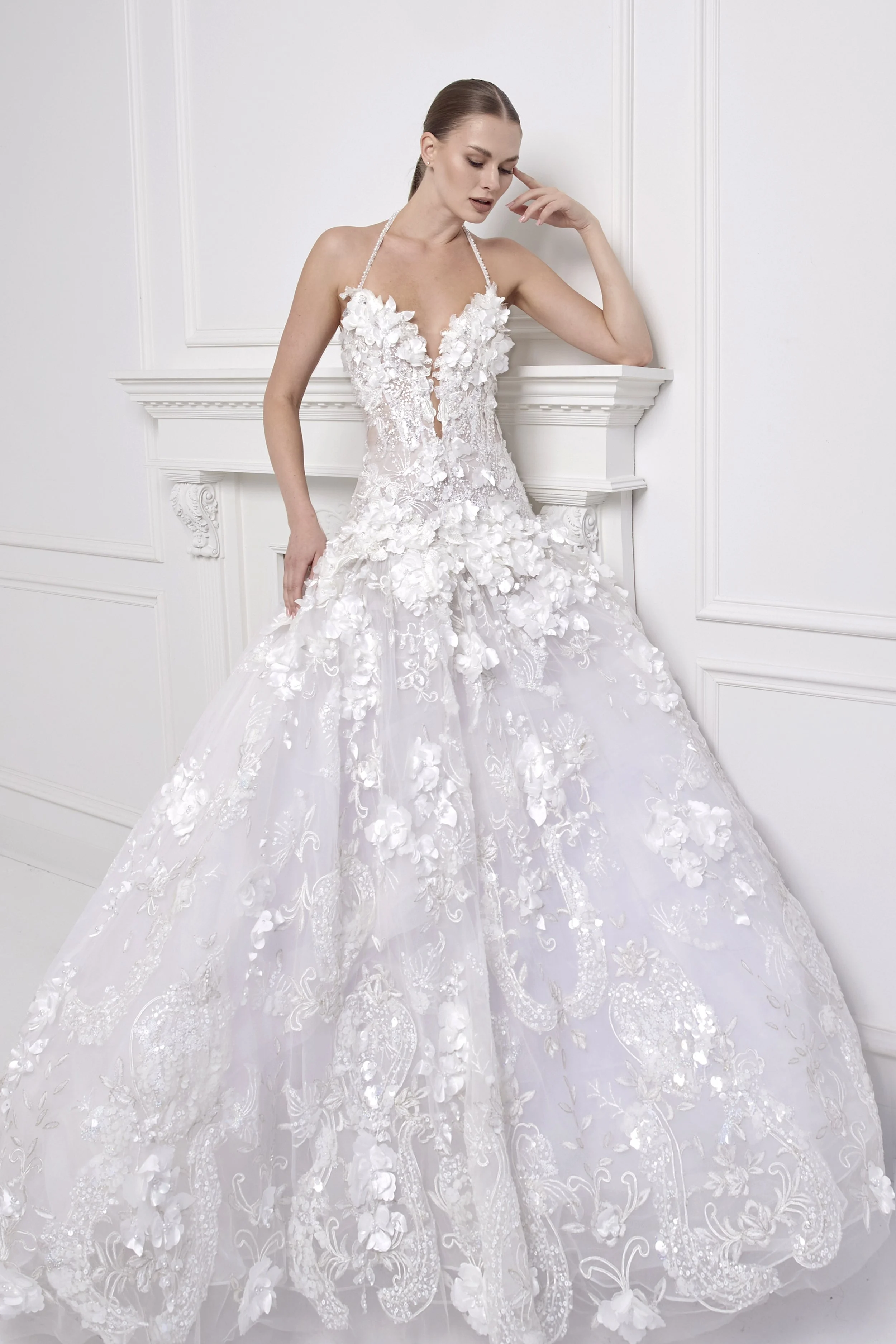 A woman in a white wedding dress with floral appliqués, standing against a white wall with decorative molding.