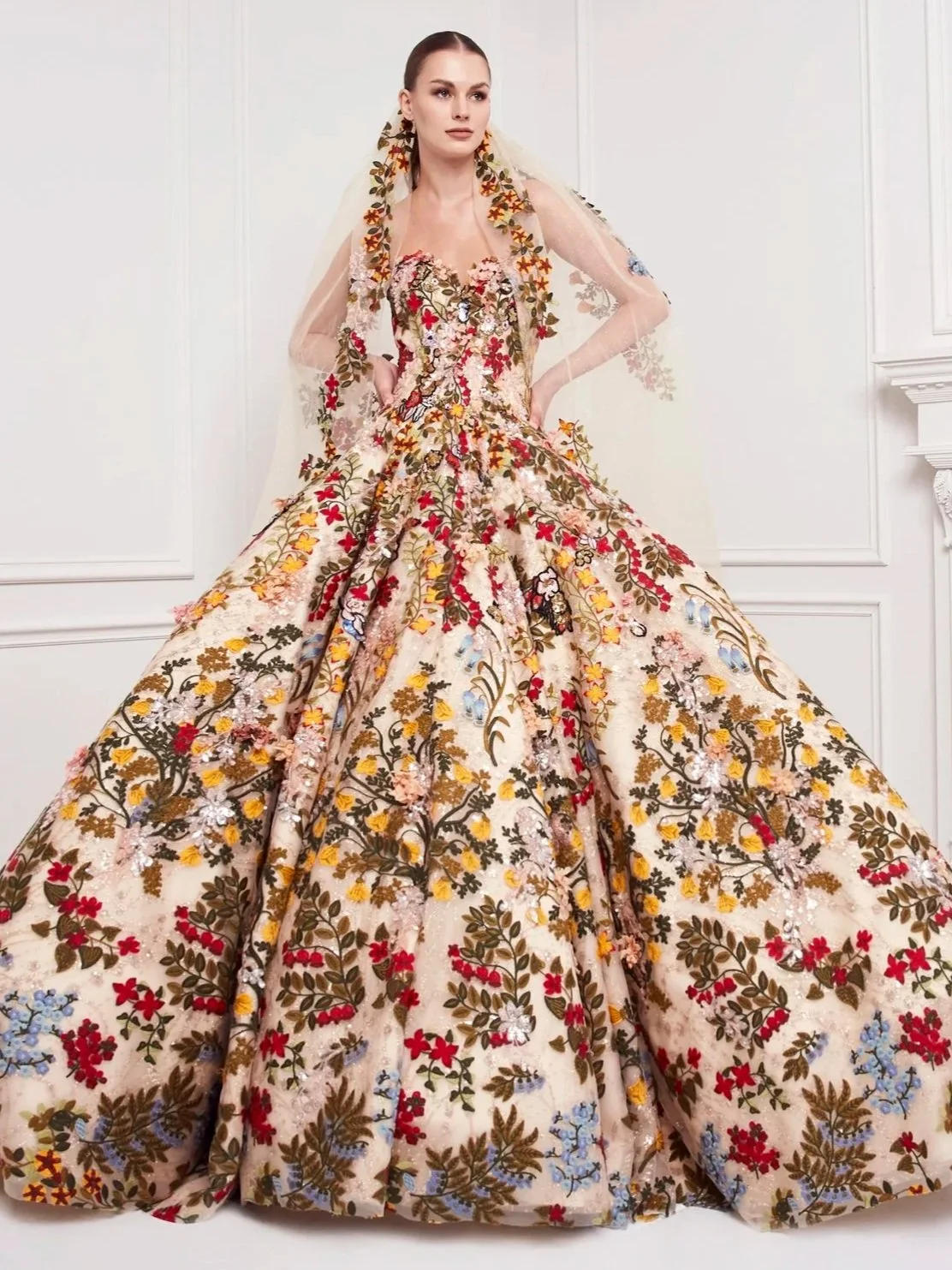 A woman modeling a floral embroidered wedding gown with a full skirt and transparent long sleeves in a white room.