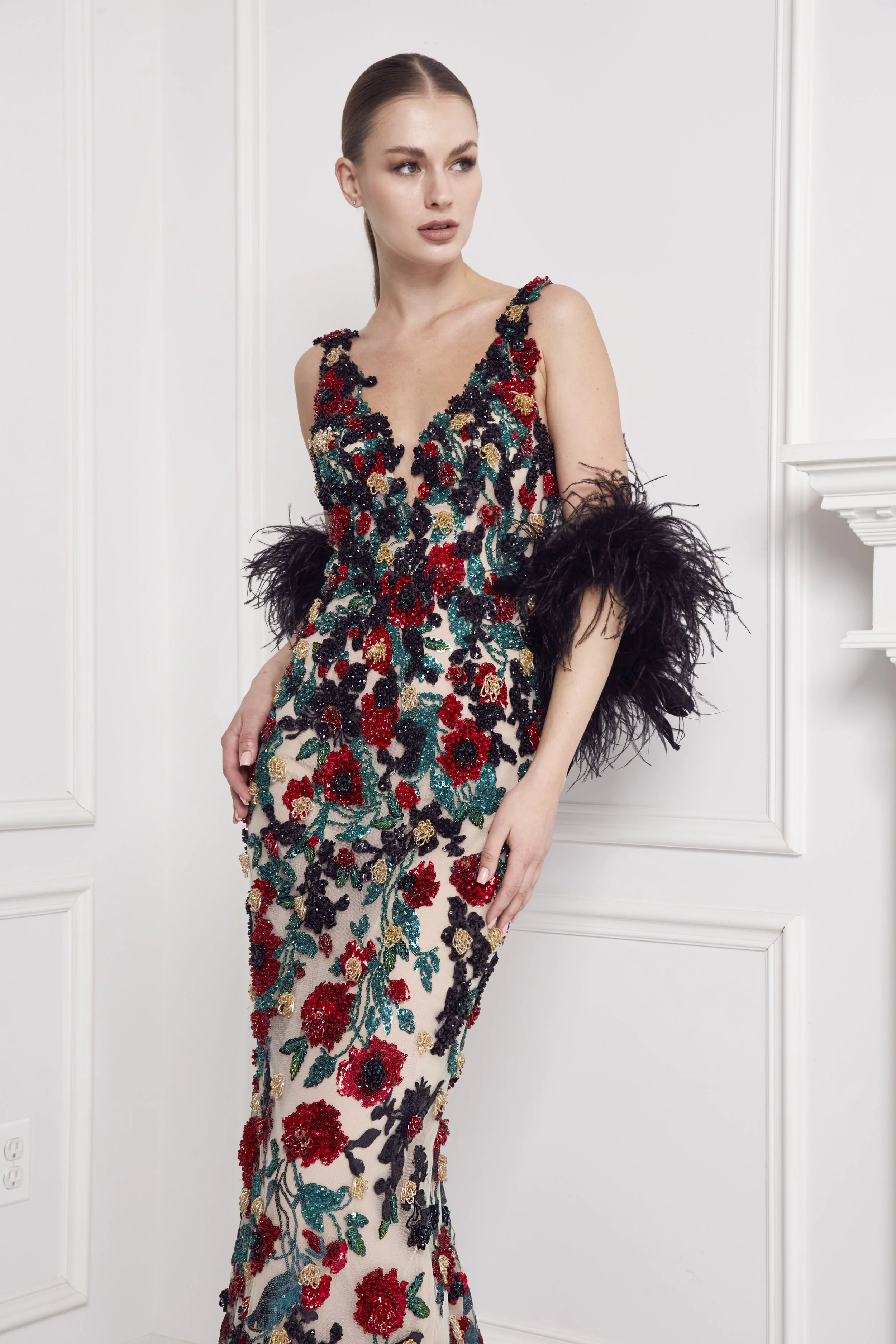 A woman wearing an elegant floor-length gown adorned with colorful floral embroidery and dark feathered accents on the sleeves, posing in a white room with decorative wall paneling.