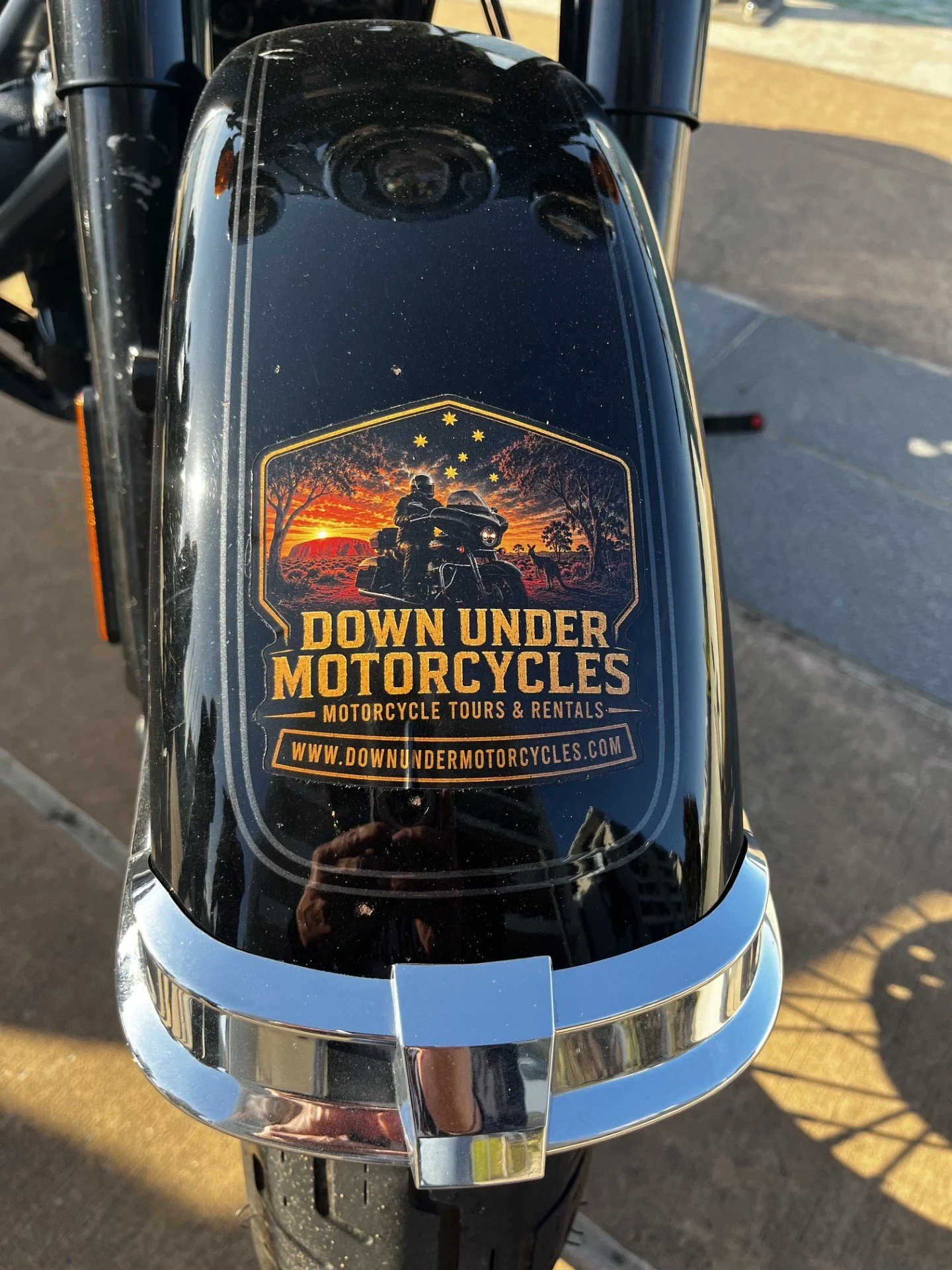 Close-up of a motorcycle fuel tank with a sticker advertising motorcycle tours and rentals, featuring a sunset scene with trees, a rider, and reindeer, and the text "Down Under Motorcycles" and "www.downundermotorcycles.com".