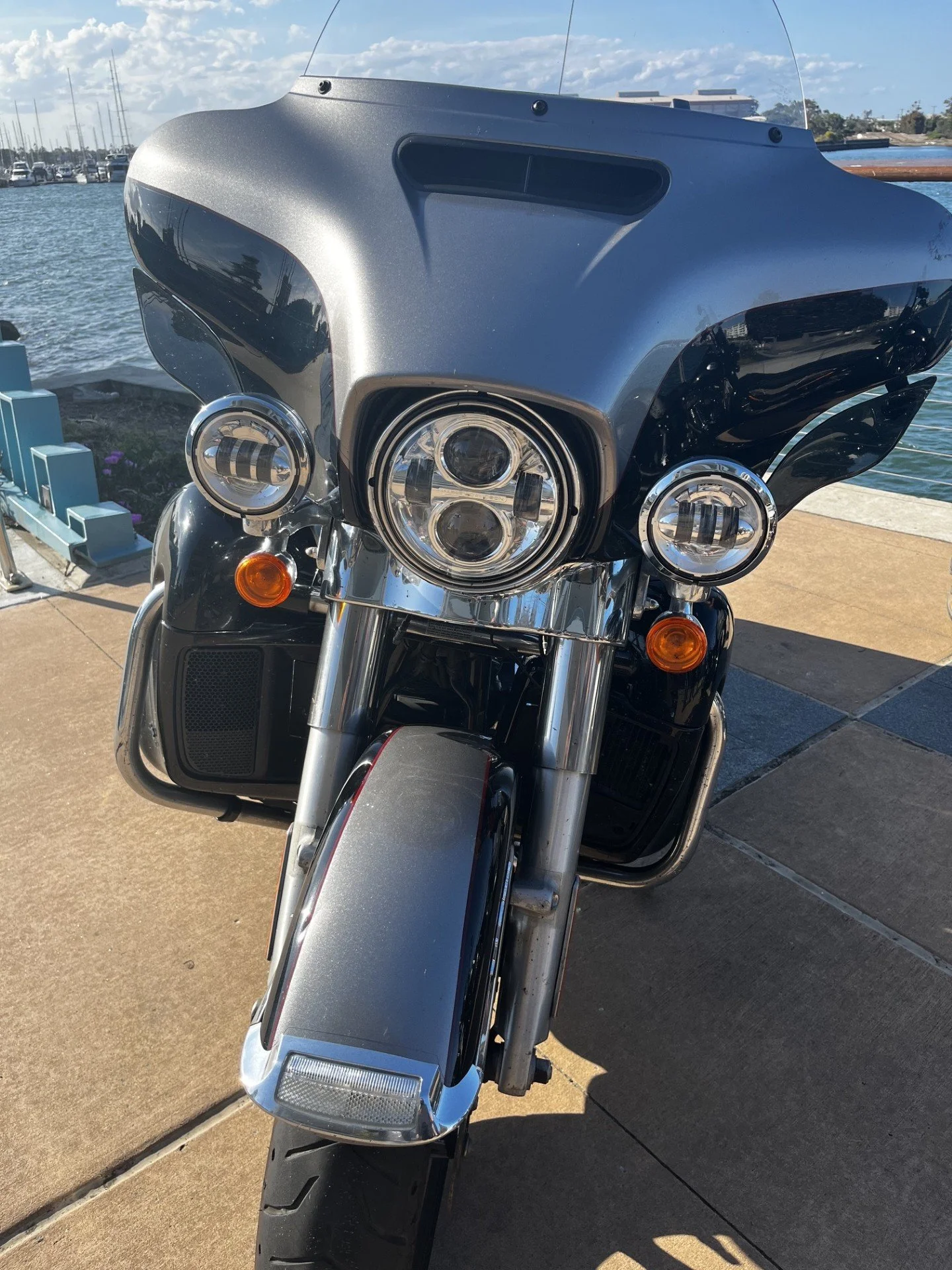 Front view of a motorcycle with headlamps, windshield, and front fork, near a body of water with boats in the background.