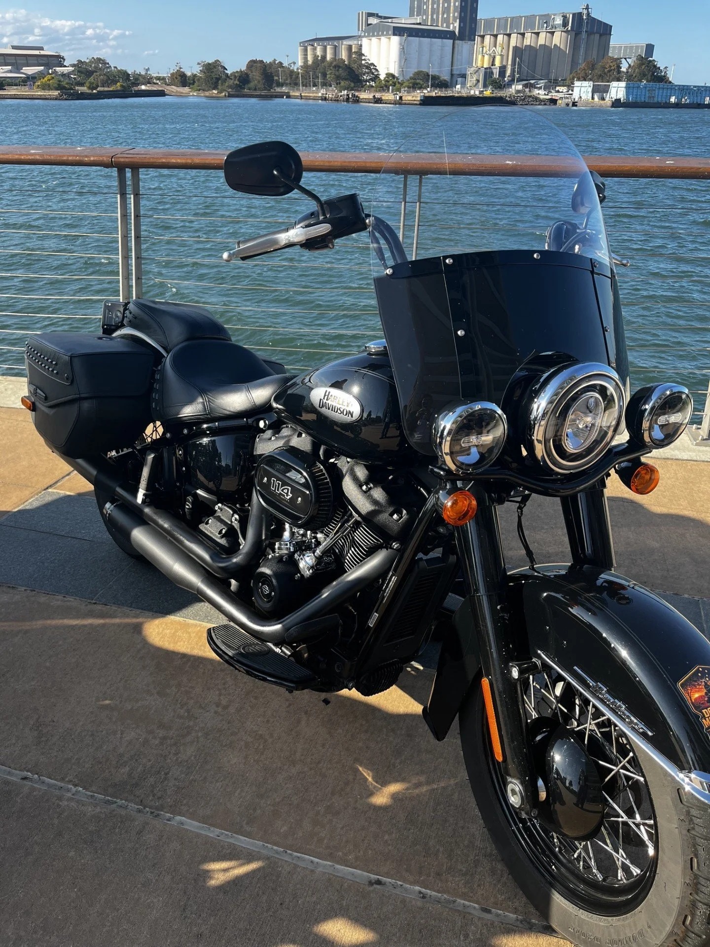 Black Harley Davidson motorcycle parked near a waterfront with industrial buildings and water in the background.