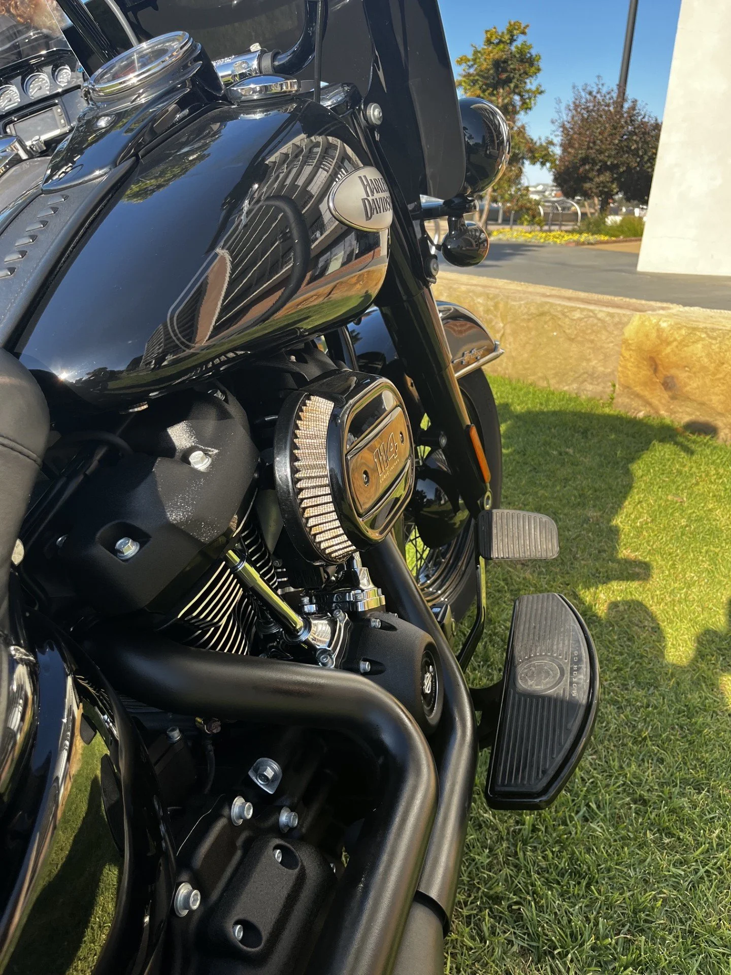 A close-up view of a black Harley-Davidson motorcycle's fuel tank, engine, and footboard, with a reflection of the building in the shiny surface of the tank, taken outdoors on a sunny day.