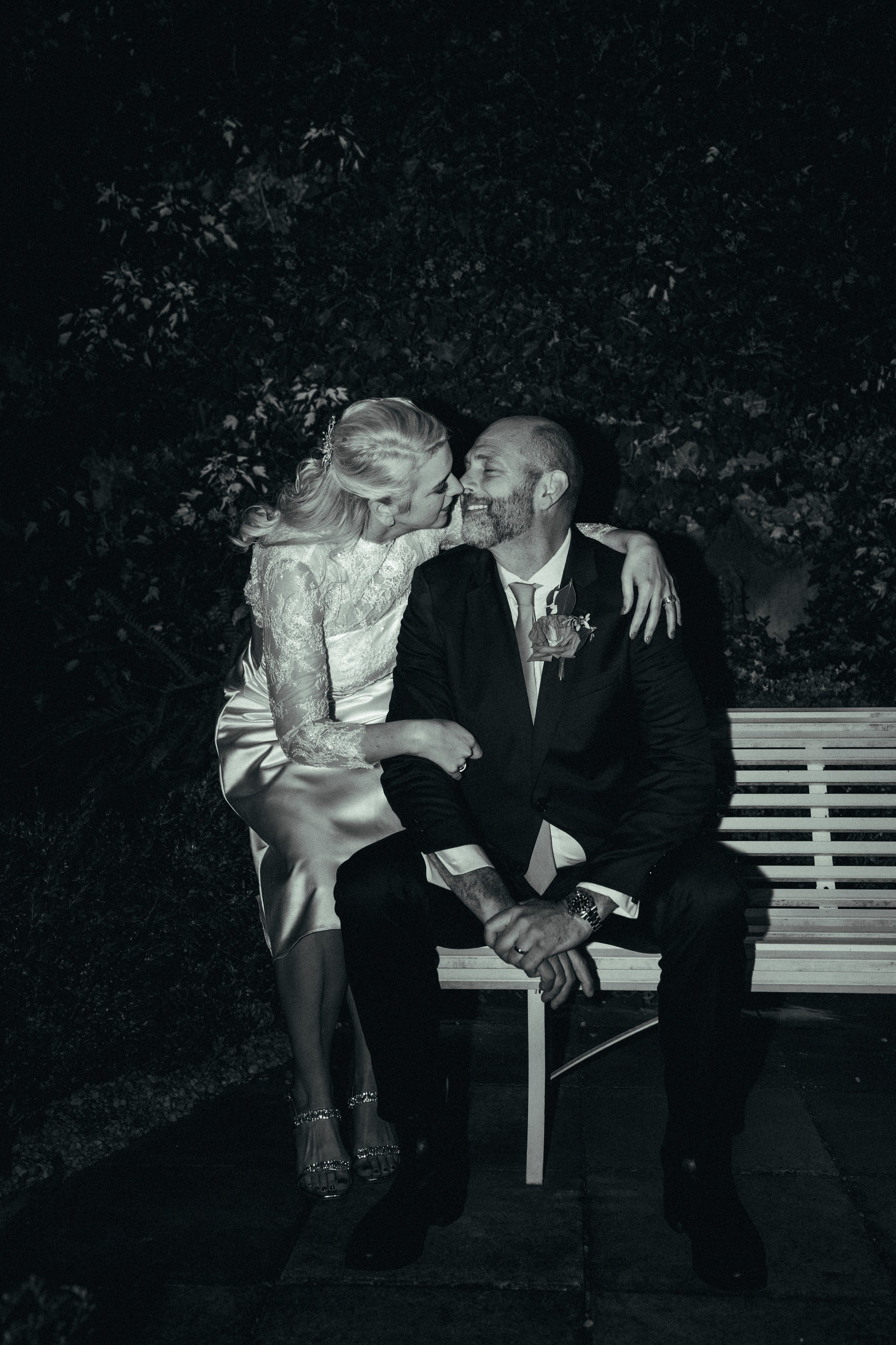 A photograph of a woman in a lace dress and high heels leaning in and about to kiss a man in a tuxedo with a boutonnière. They are sitting on a bench outside at night, surrounded by foliage.