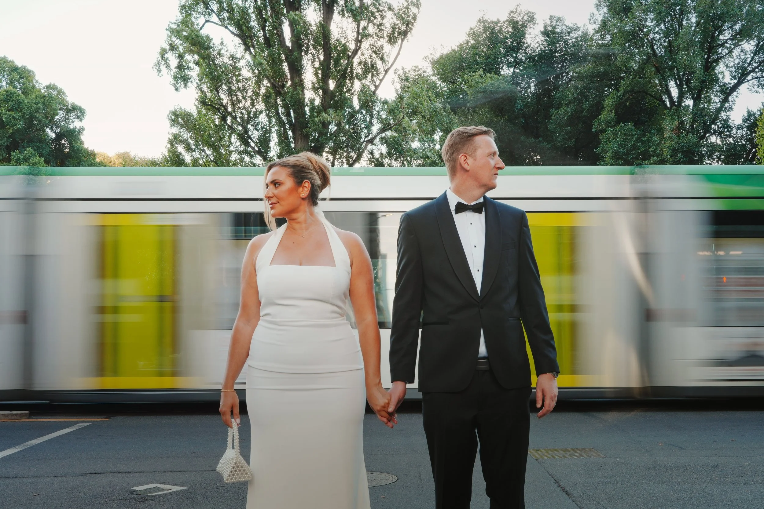 A bride and groom holding hands while standing in front of a moving train with trees in the background.