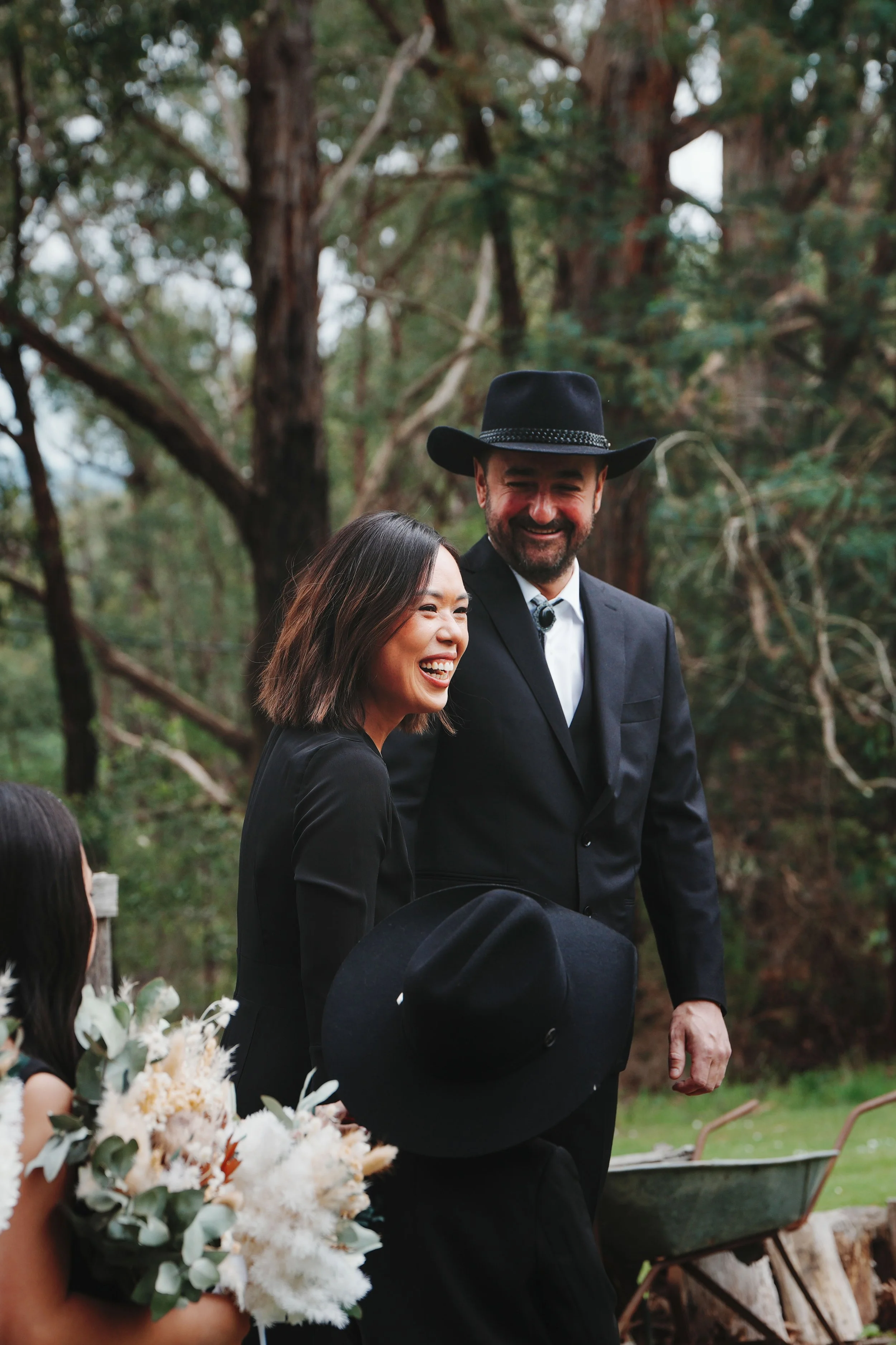 A man wearing a black cowboy hat and a woman with shoulder-length dark hair are smiling outdoors near trees, possibly at a garden or outdoor event. The woman is holding a bouquet of white and green flowers.