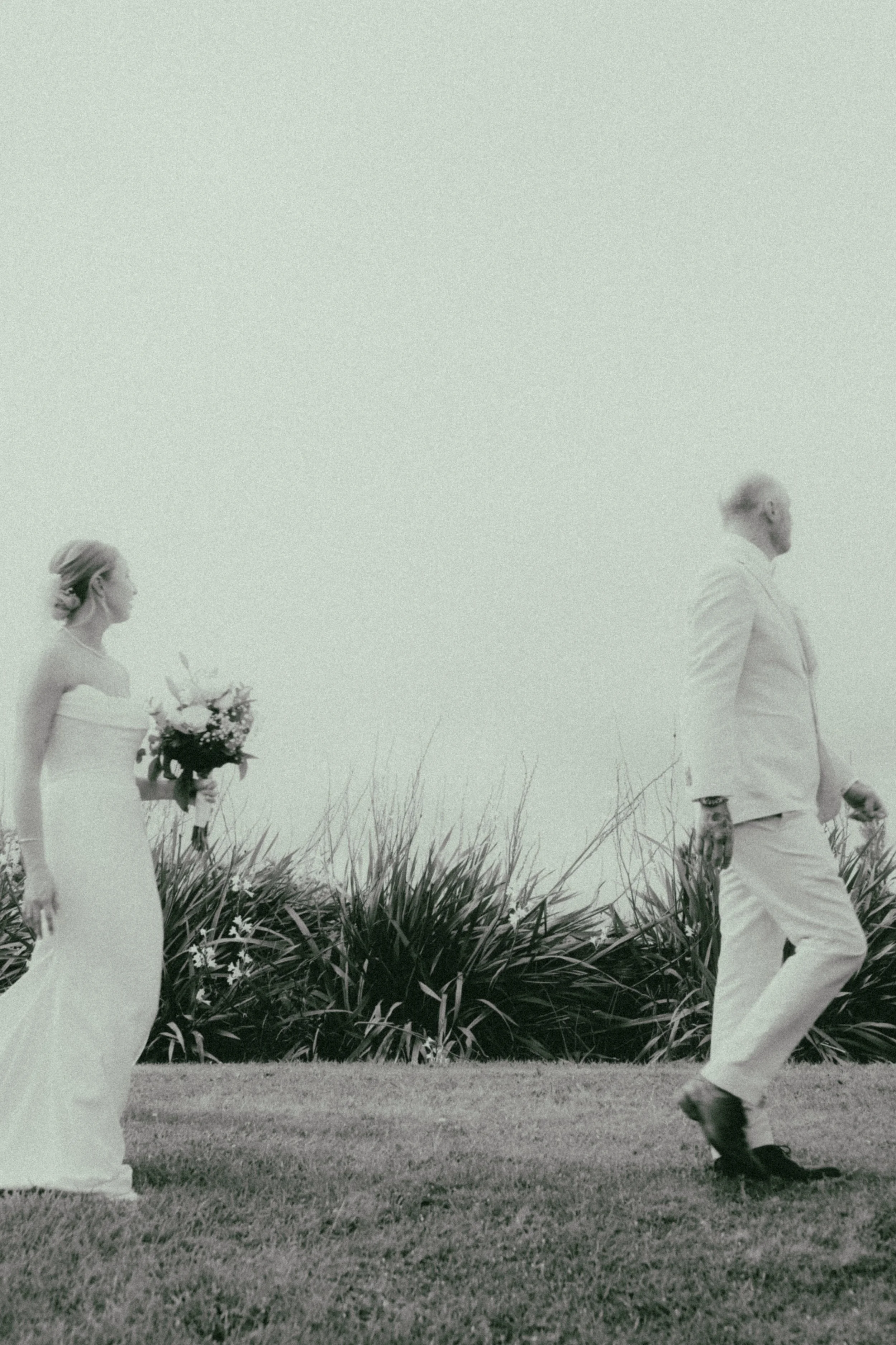 A black and white photo of a bride in a white wedding dress holding a bouquet of flowers, and a groom in a white suit walking outdoors on grassy terrain with tall plants in the background.