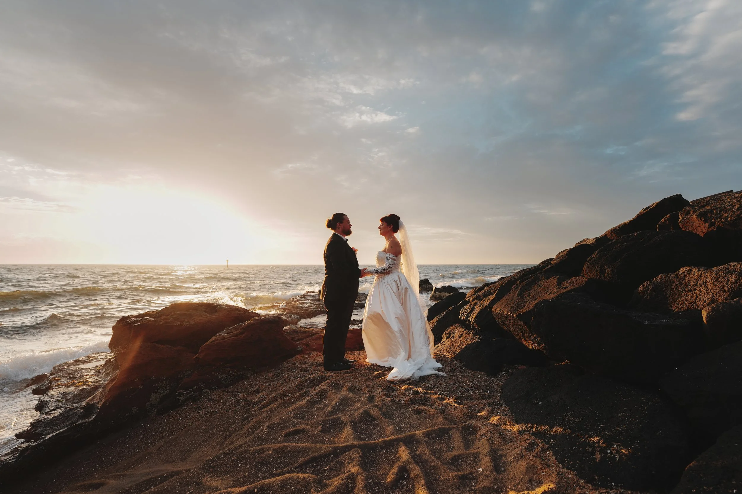 A bride and groom holding hands on the rocks by the beach at sunset, with the ocean and cloudy sky in the background.