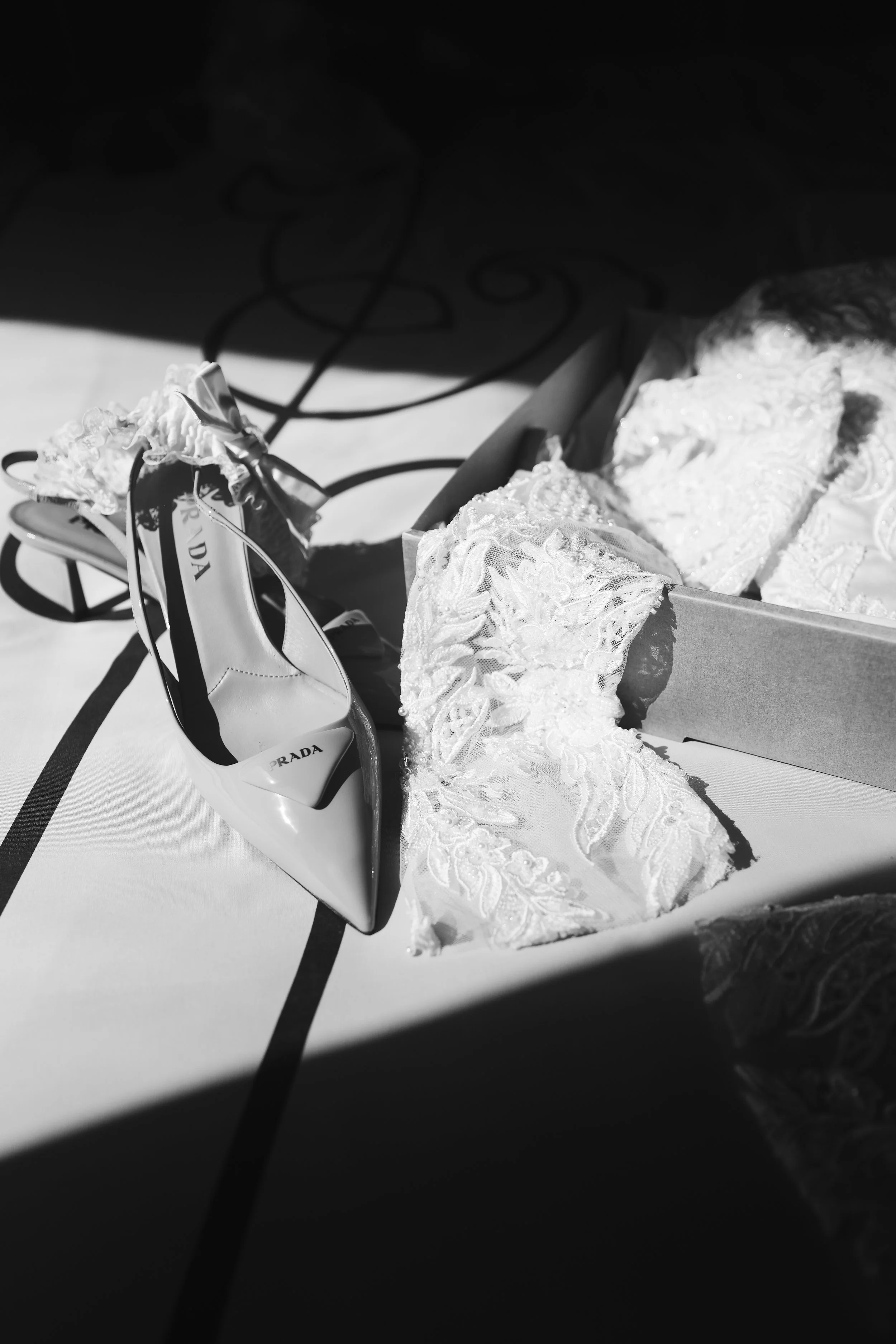 Black and white photo of lace wedding dress, a pair of white high heels with brand 'Prada' written on the insole, placed on a table with a box or tray containing lace fabric.