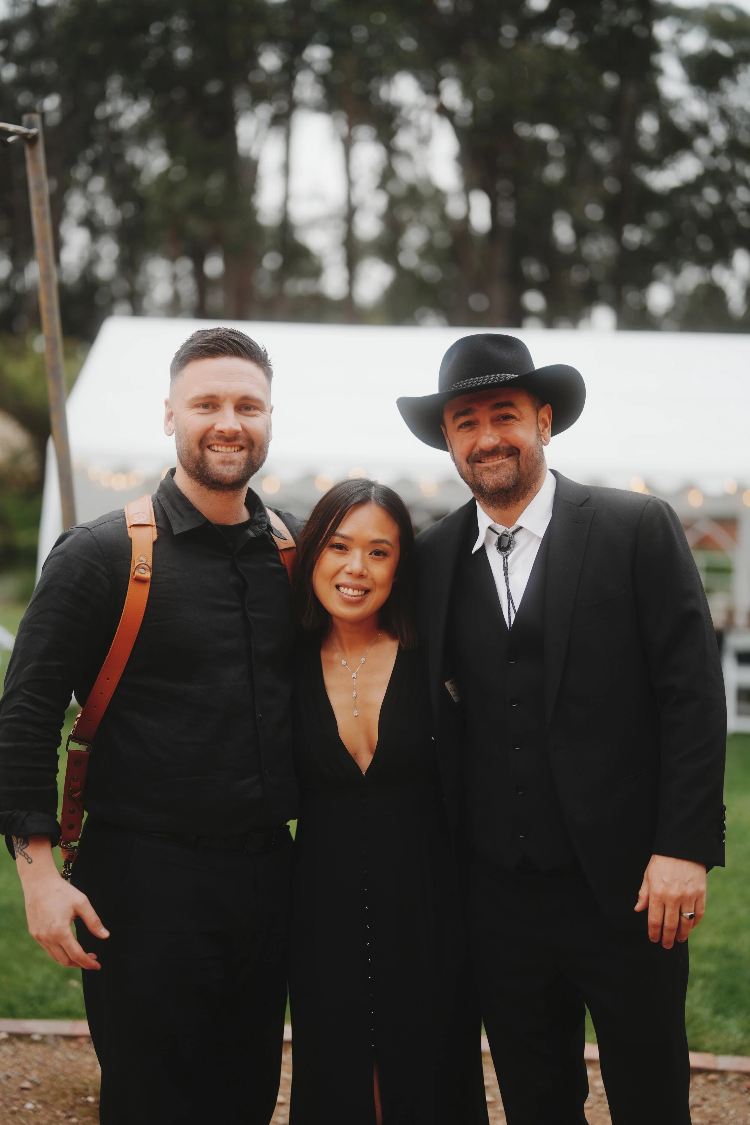 Three people smiling outdoors at a gathering, with trees and a white tent in the background. The person on the left is a man with short hair, a beard, wearing a black shirt and a brown strap, possibly from a bag. The person in the middle is a woman with shoulder-length dark hair, wearing a black top with a deep neckline, and a necklace. The person on the right is a man with a beard, wearing a black cowboy hat, a black suit with a white shirt, and a bolo tie.