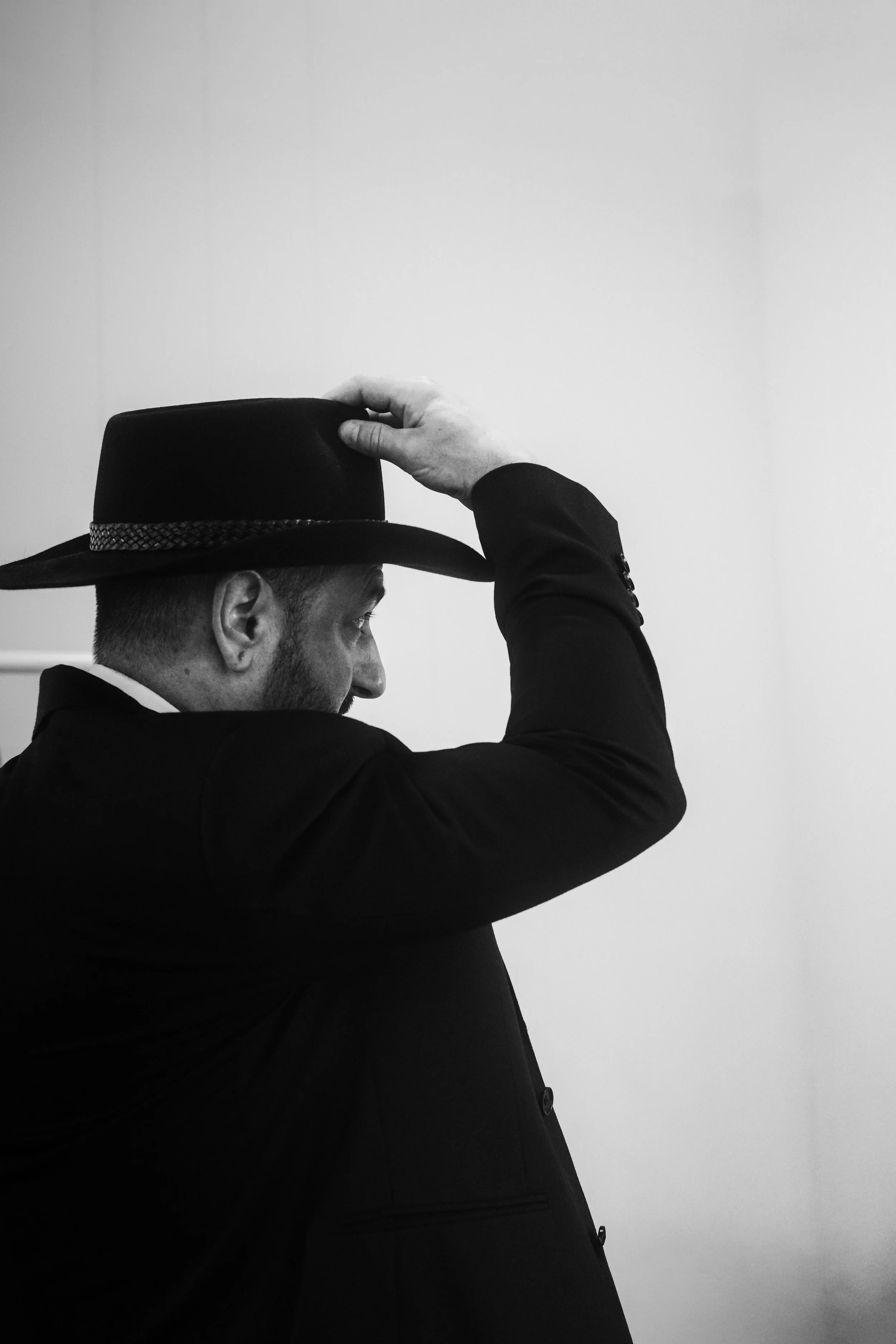 A man dressed in a dark suit, white shirt, is adjusting his black fedora hat with a woven band, standing against a plain white wall.