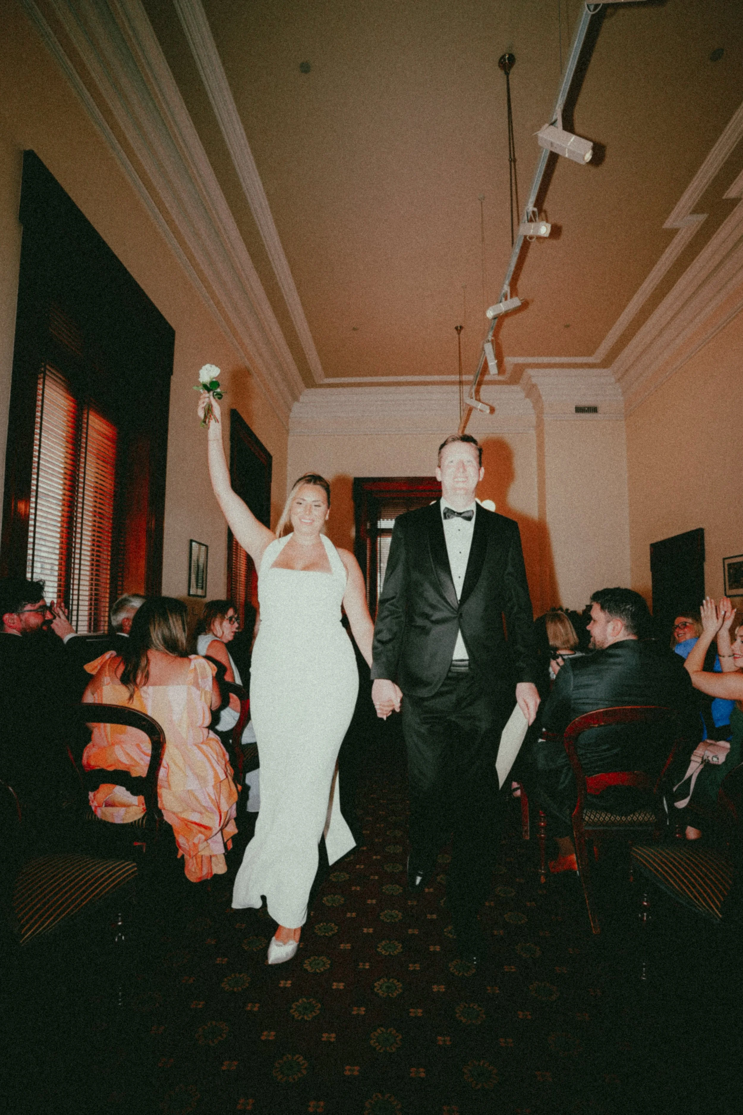A bride in a white gown holding a flower and a groom in a tuxedo holding hands, walking down an aisle at a wedding reception surrounded by seated guests.