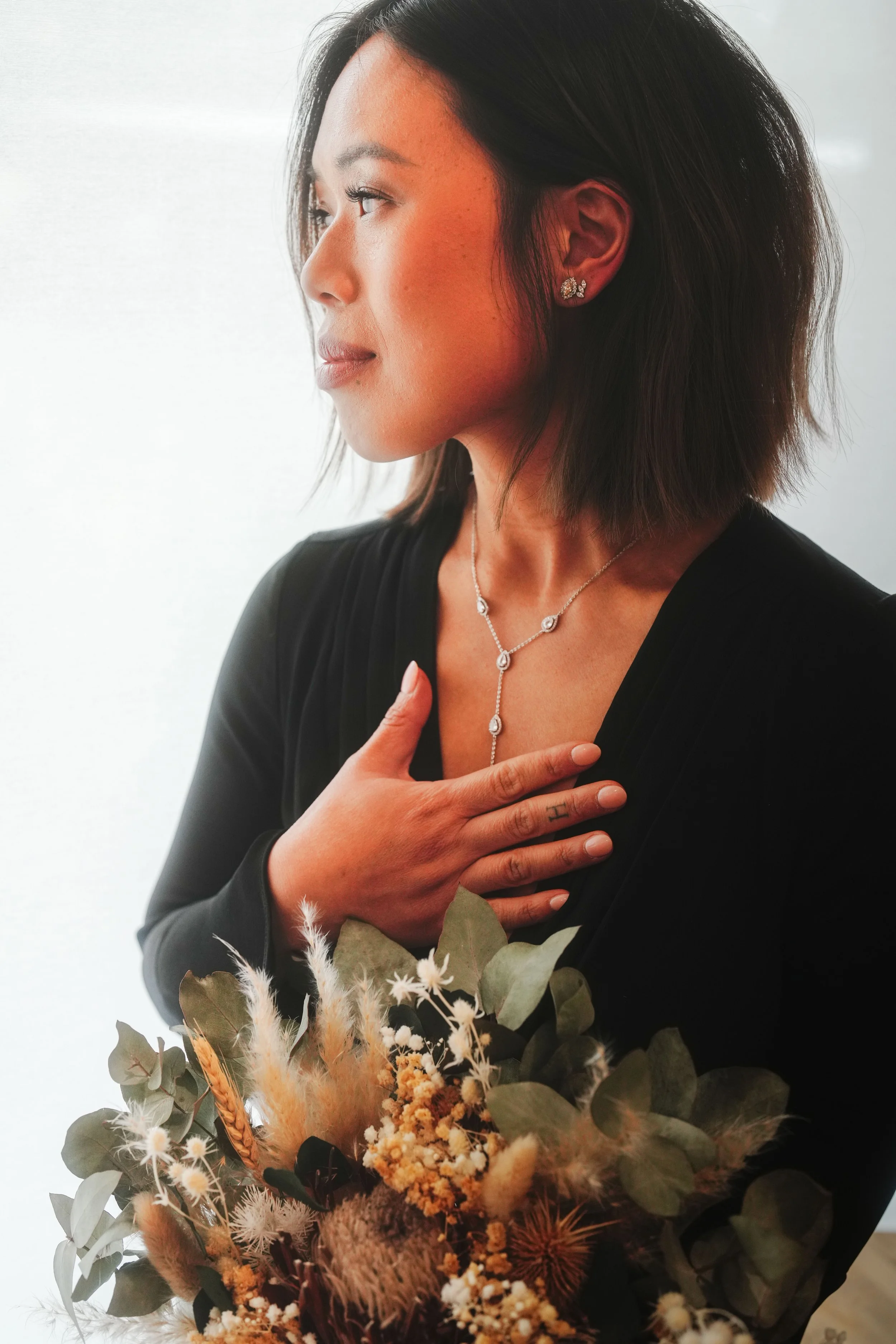 A woman with dark hair wearing earrings and a necklace, holding a bouquet of dried flowers and greenery, with her hand on her chest.