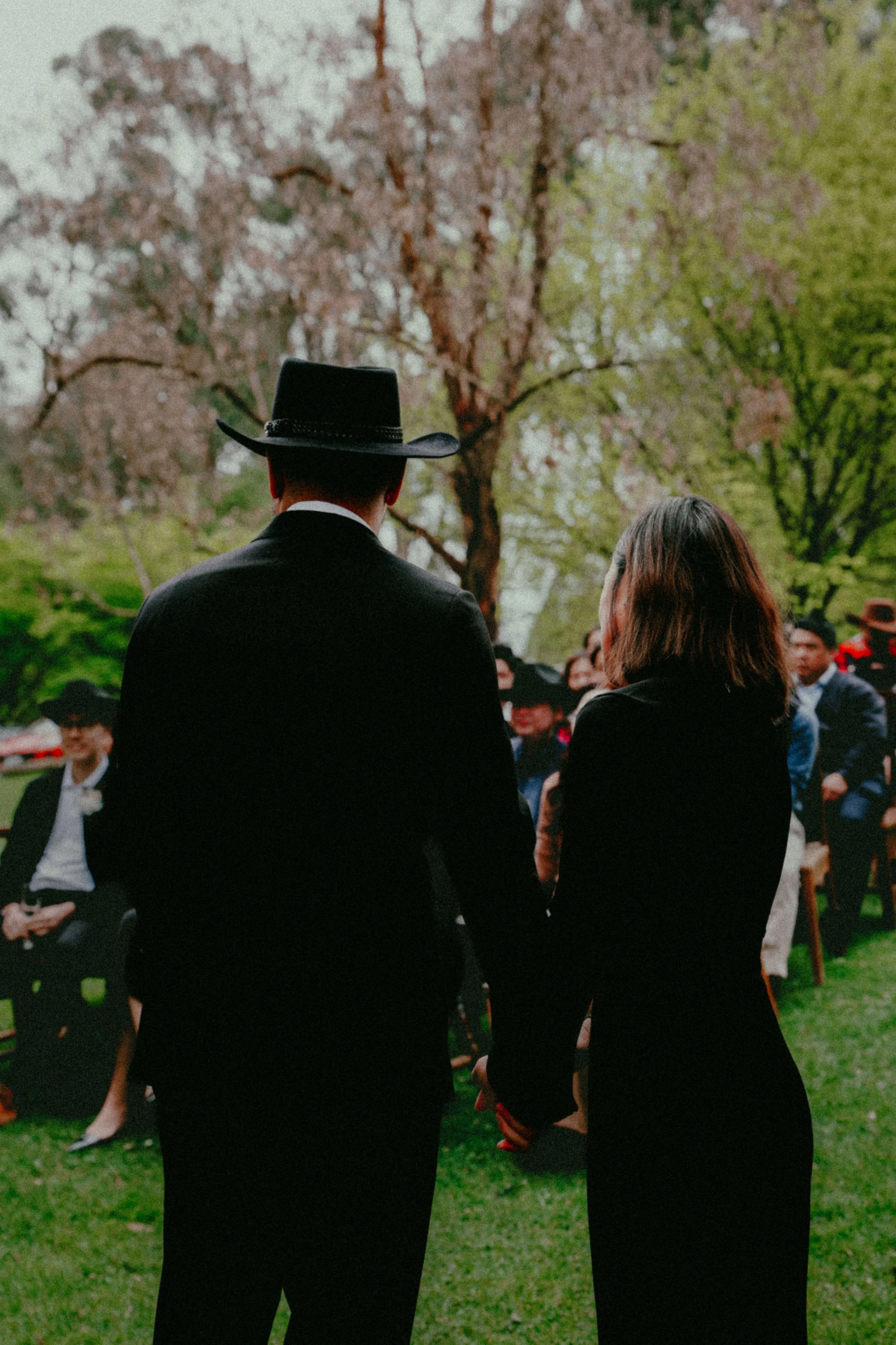 A man and a woman standing outdoors in front of blossoming trees, with a crowd of people in the background.