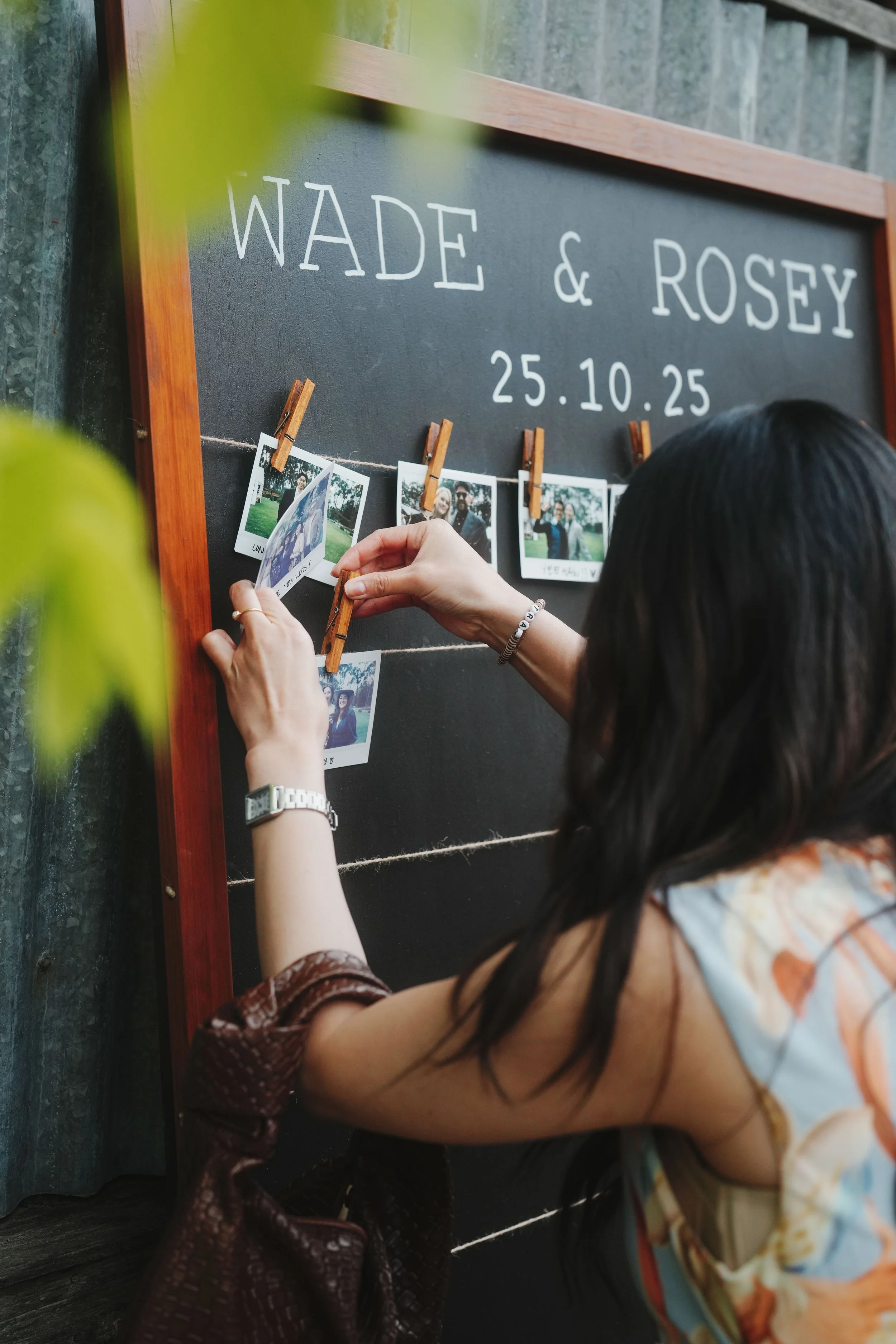 Person hanging photographs on a black display board labeled 'WADE & ROSEY' with date '25.10.25'
