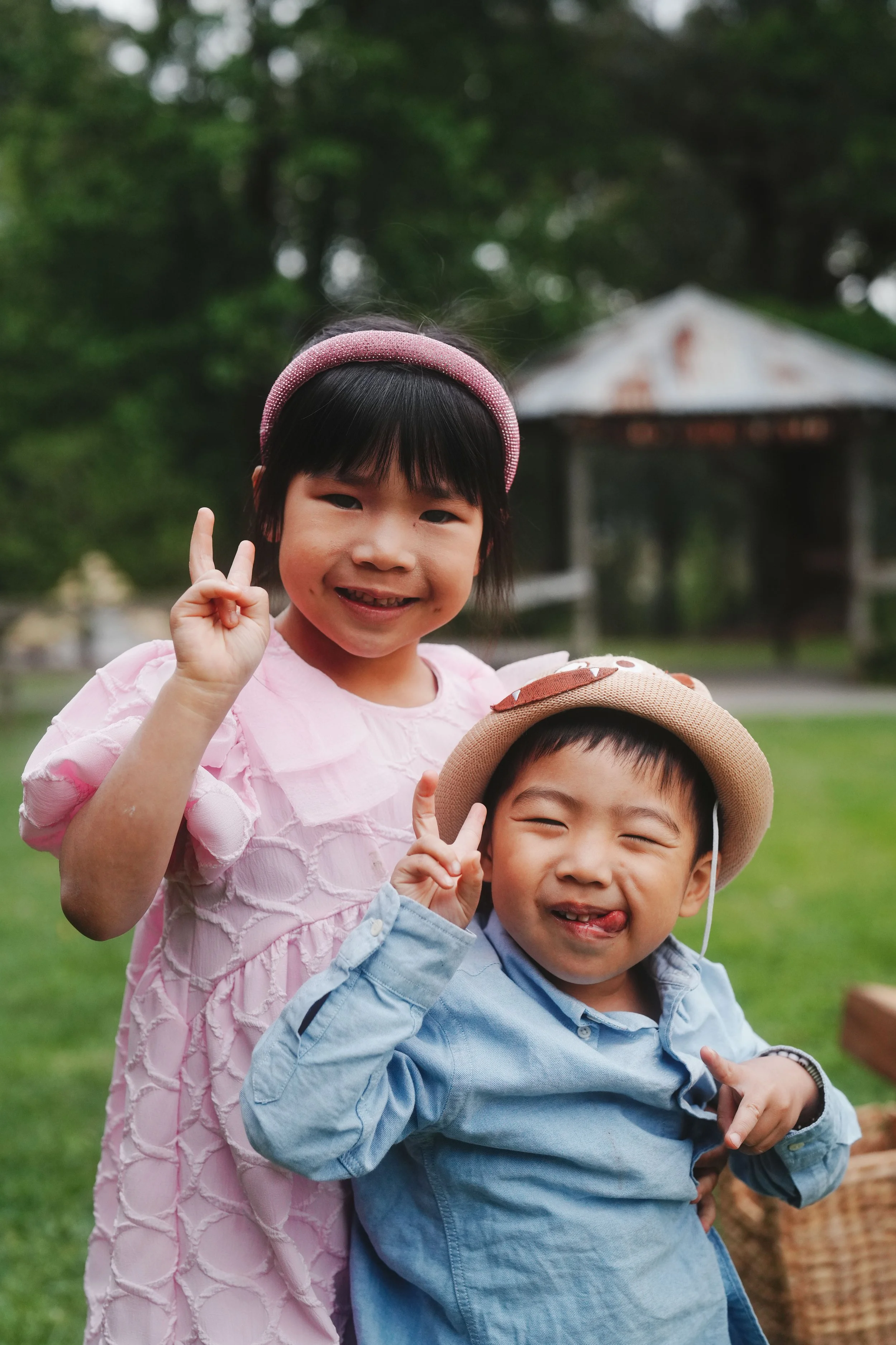 Two smiling children, a girl and a boy, outdoors in a park, making peace signs with their hands. The girl has short black hair, wearing a pink dress and headband. The boy has short dark hair, wearing a light blue shirt and a straw hat. Behind them is