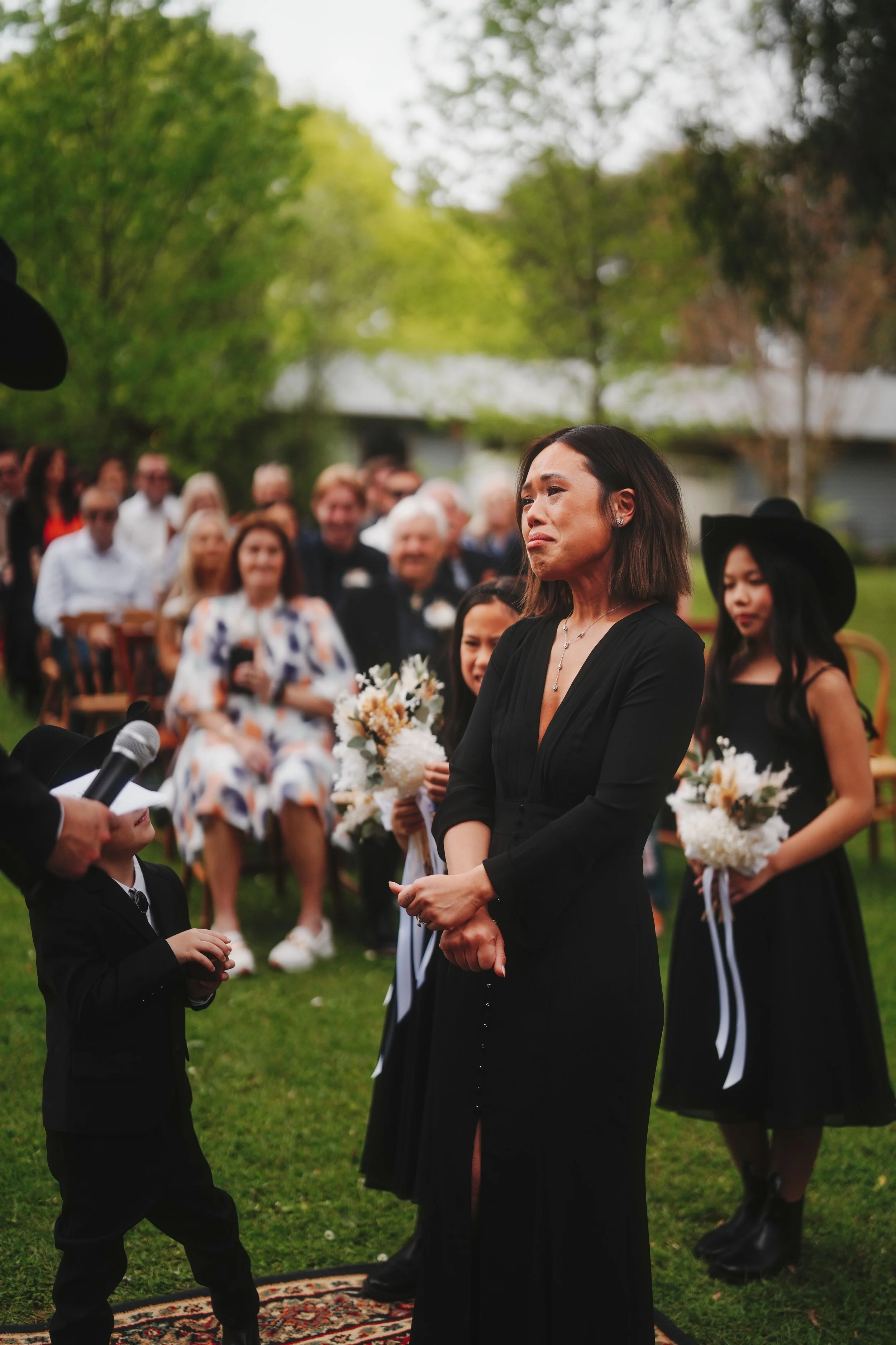 A woman in a black dress appears emotional at an outdoor event, with a crowd gathered behind her on a lawn with trees.