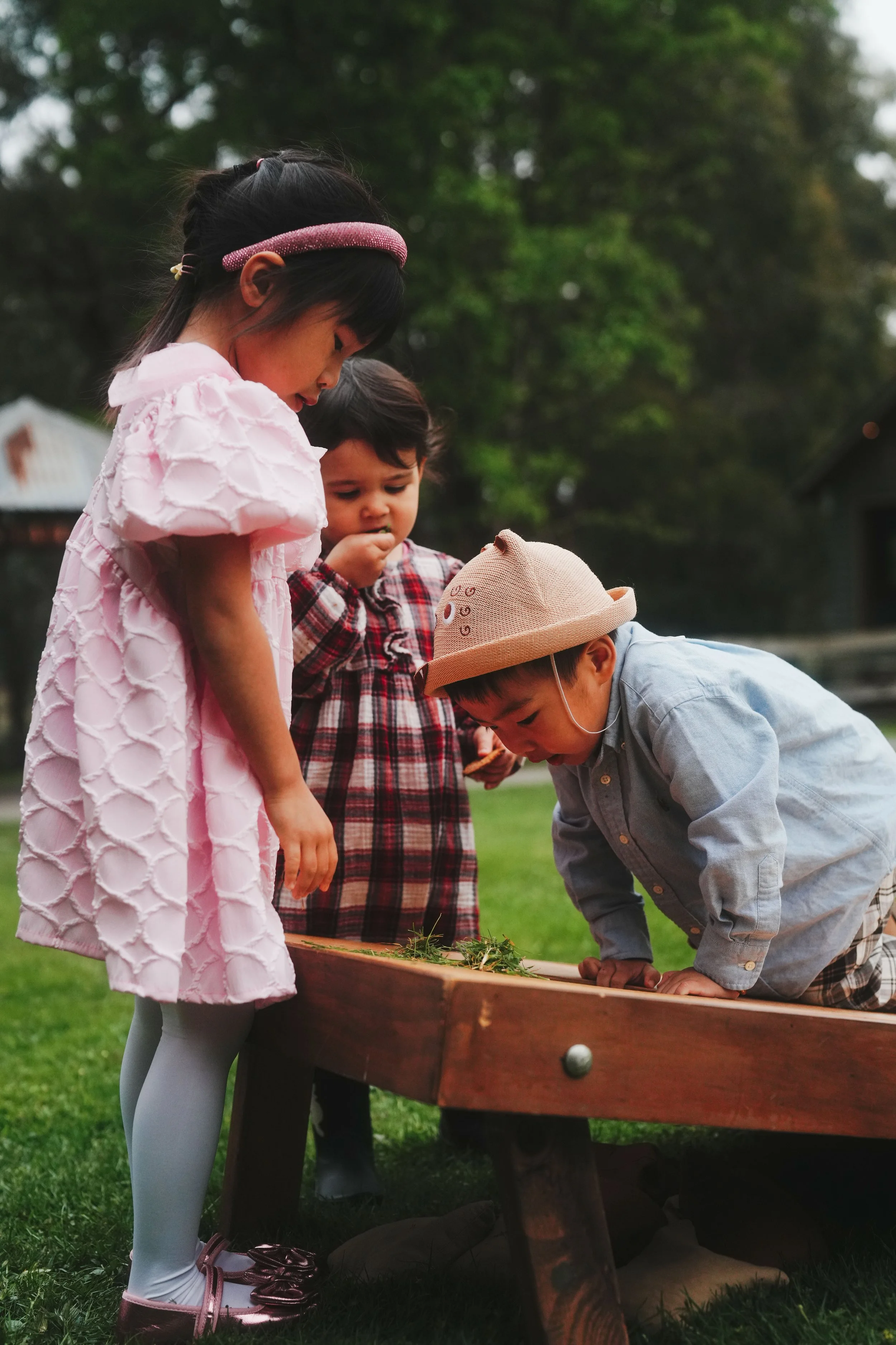 Three children gathered around a game table outdoors, looking at a game board with greenery and trees in the background.