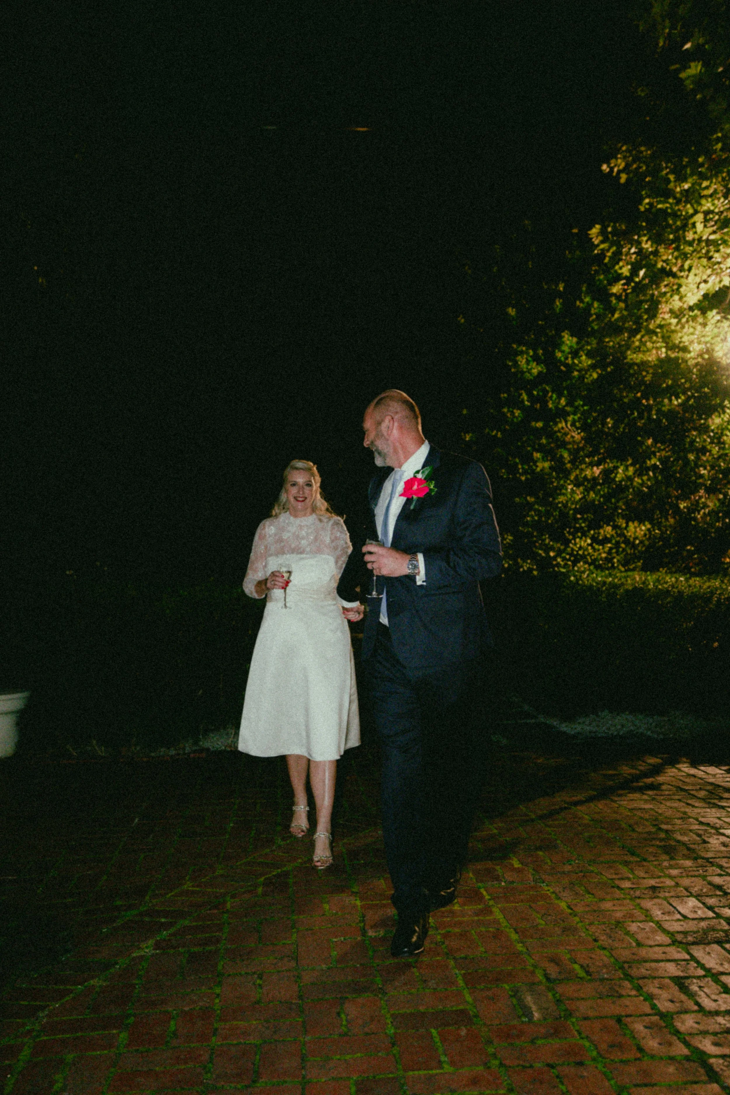 A bride in a white lace dress holding a glass and smiling, talking to a man in a dark suit with a red rose boutonniere, at night under trees with lit lights.