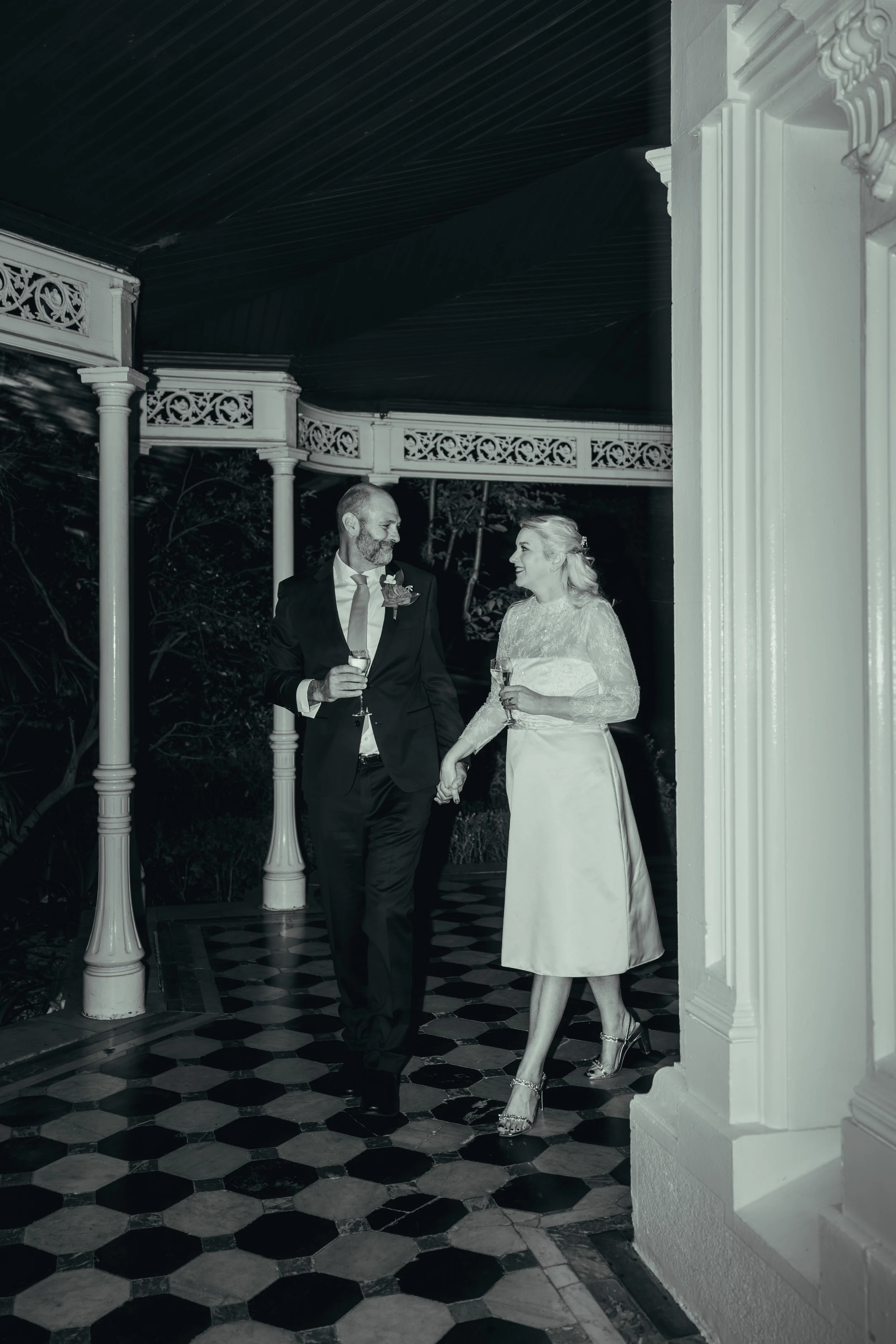A bride and groom in wedding attire holding hands and each holding a glass, smiling at each other, standing indoors with decorative pillars and patterned floor tiles.
