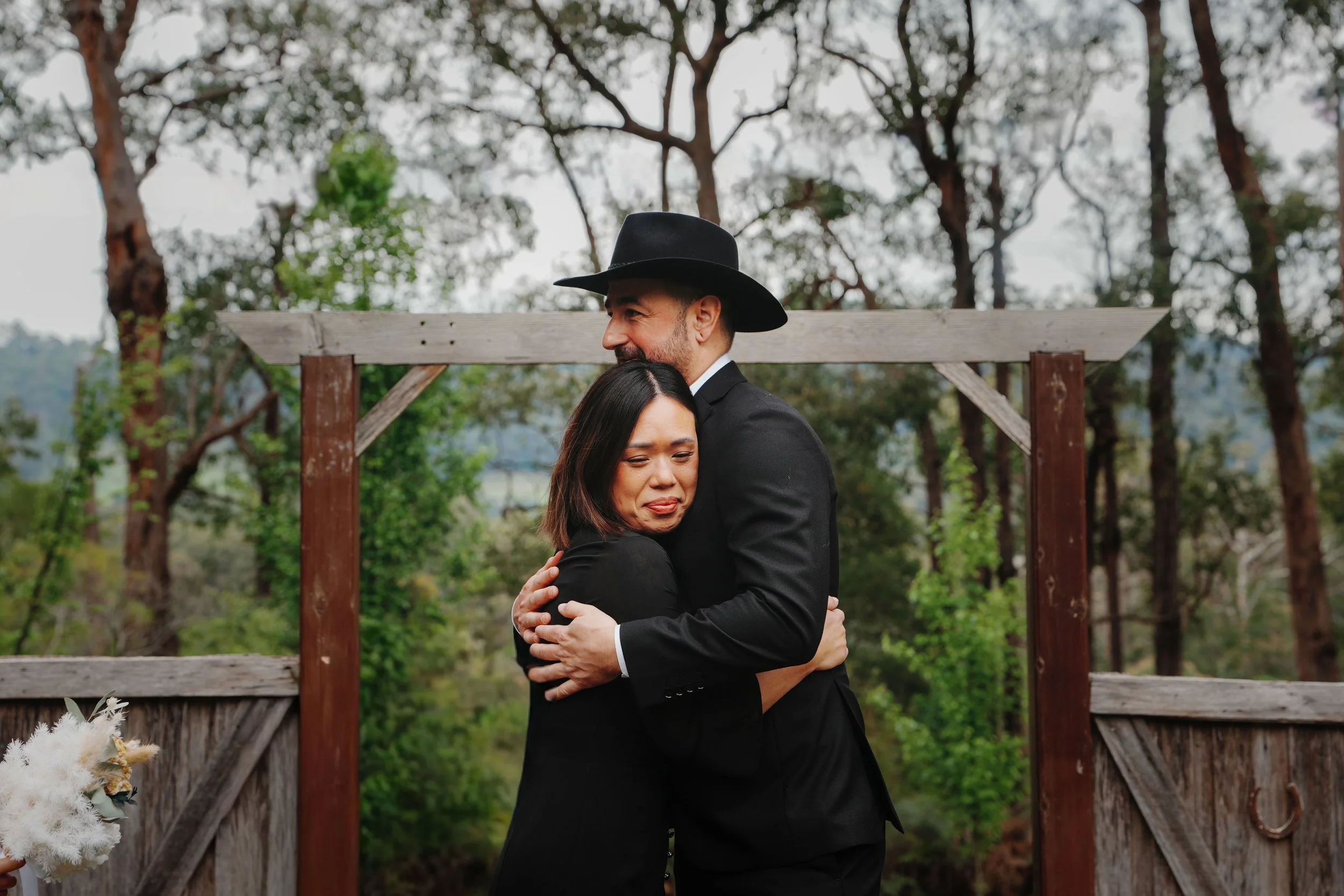 A man and woman hugging outdoors in a wooded area on a cloudy day, with a wooden fence in the background.