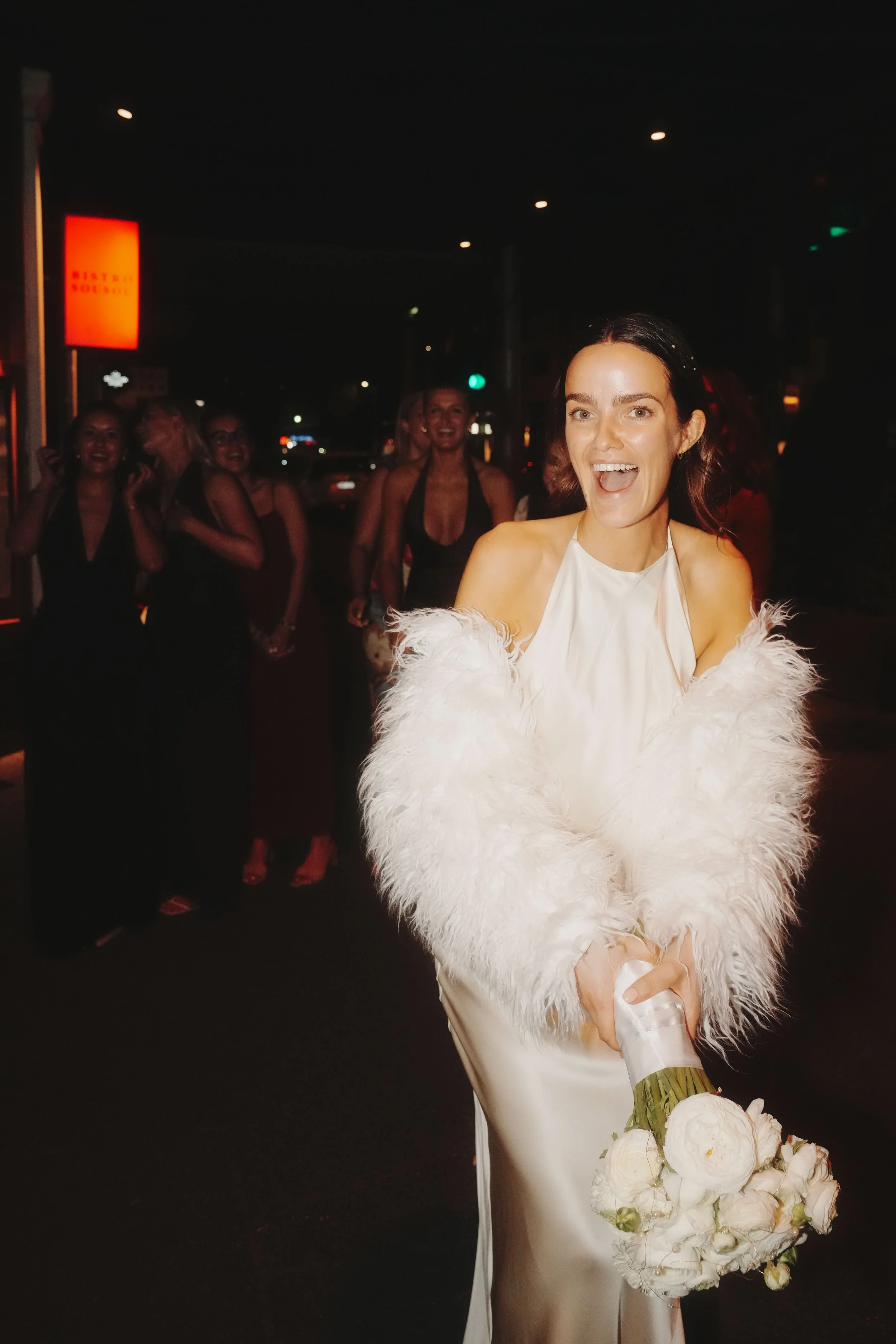 A woman in a white dress with a fur stole holding a bouquet of white flowers on her wedding day, smiling with friends in the background at night.