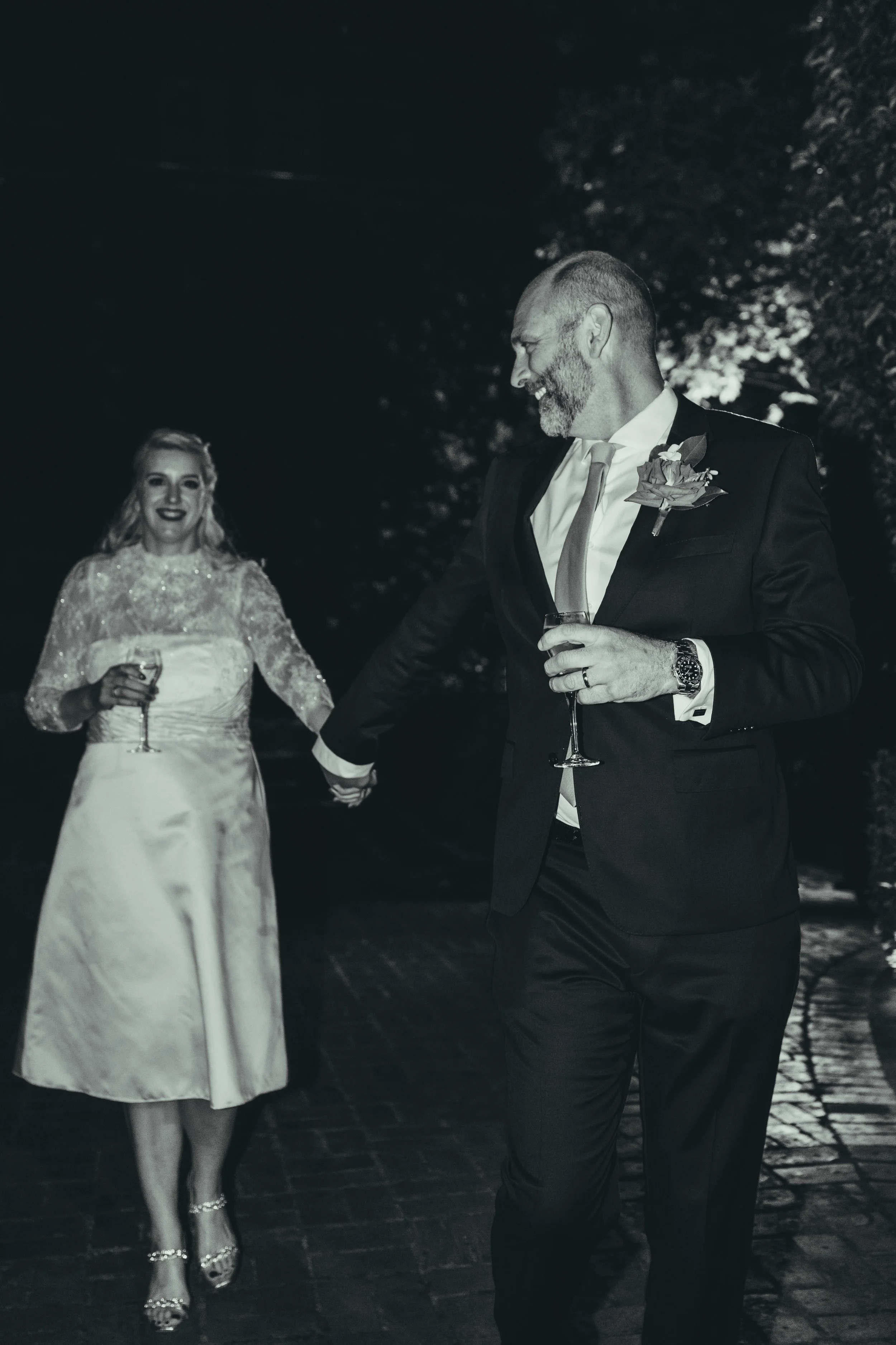 A man and a woman are walking hand in hand outdoors at night, both holding champagne glasses and smiling. The woman wears a white dress with lace details, and the man is in a black suit with a boutonniere, tie, and watch.