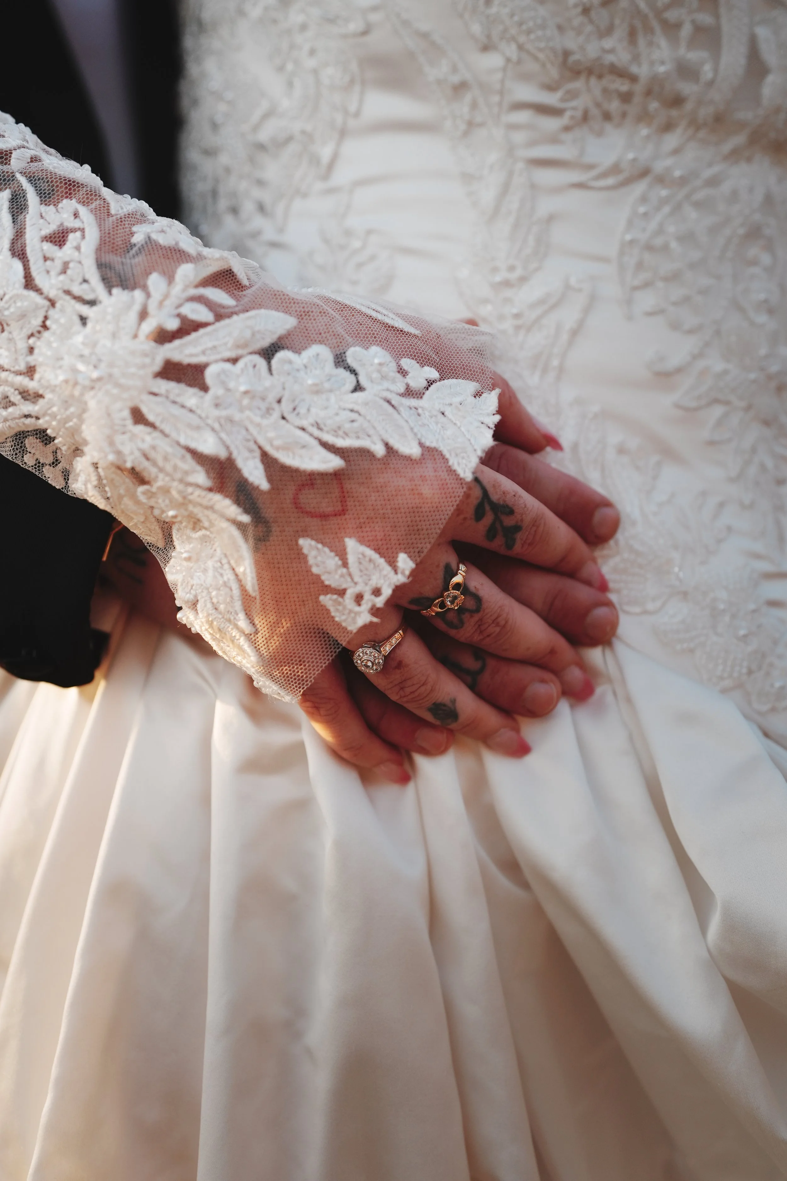 Close-up of a person's hand resting on a wedding dress. The hand has tattoos and is wearing rings. The wedding dress features intricate lace detailing on the sleeve and a satin skirt.