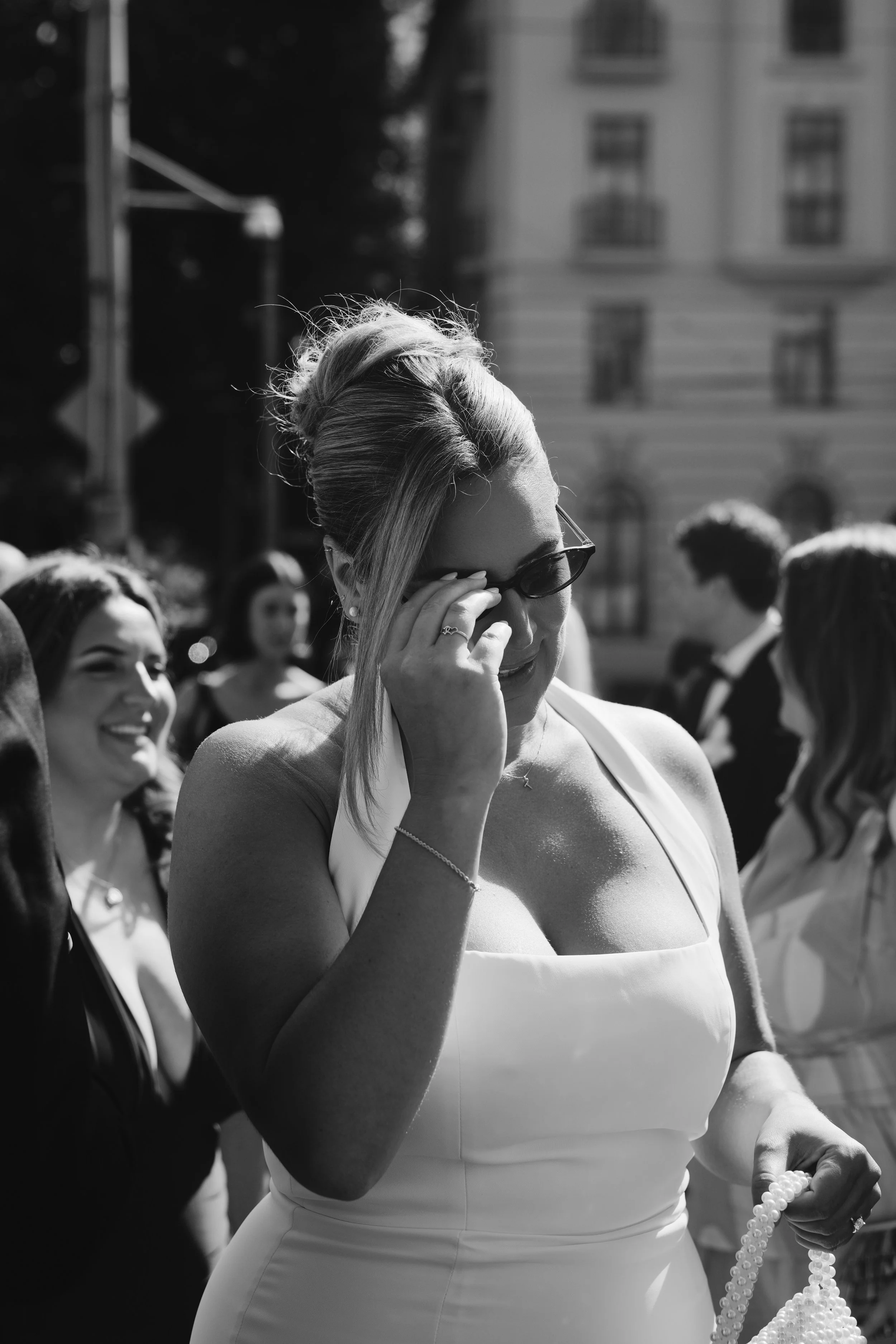 A black and white photo of a woman wearing sunglasses and a sleeveless dress, holding a pearl necklace, surrounded by people outdoors, with a building in the background.