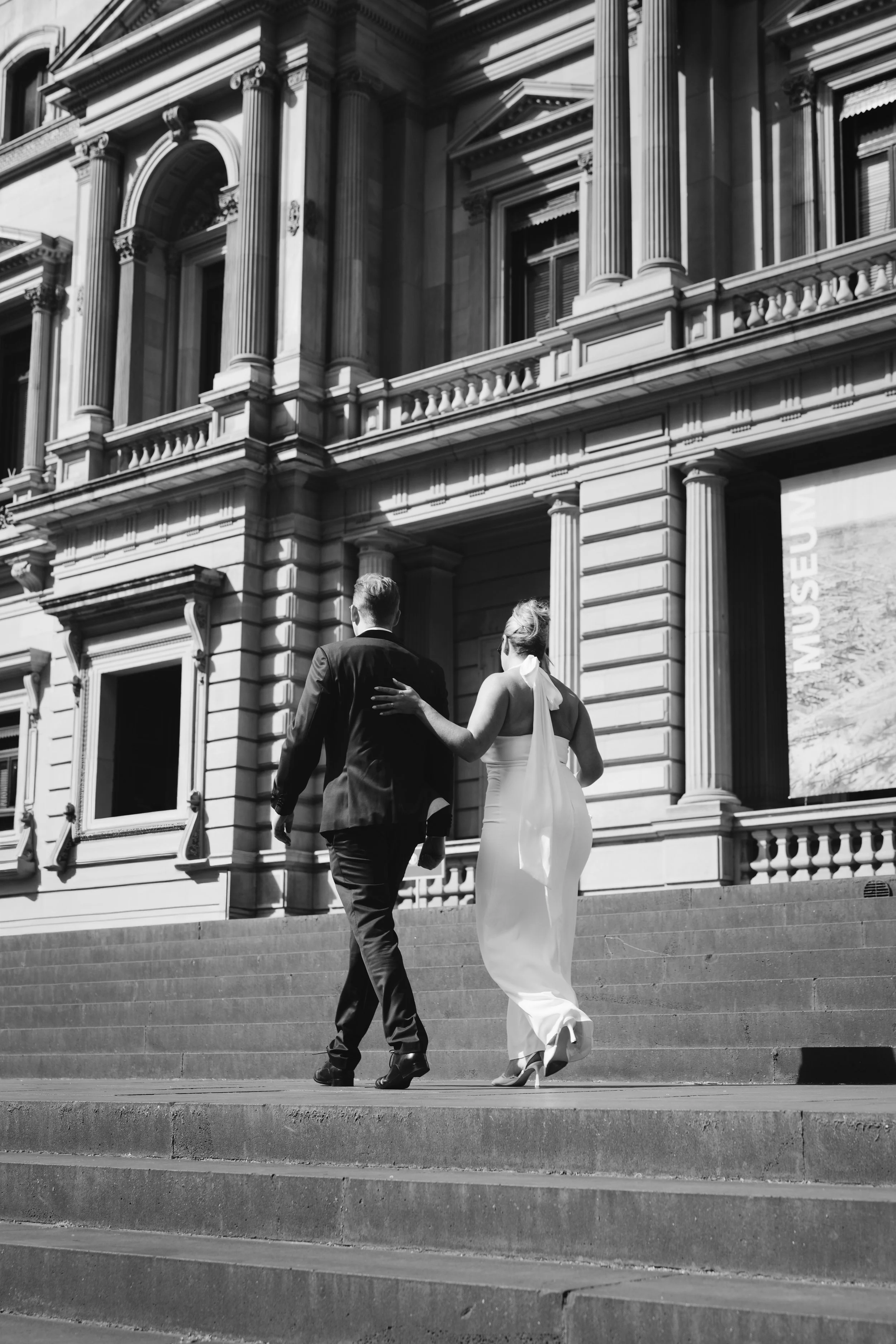 A black-and-white photo of a man and woman walking up outdoor stairs in front of a grand, historic building with columns and ornate architectural details. The man is wearing a suit, and the woman is wearing a long dress with a bow around her neck.
