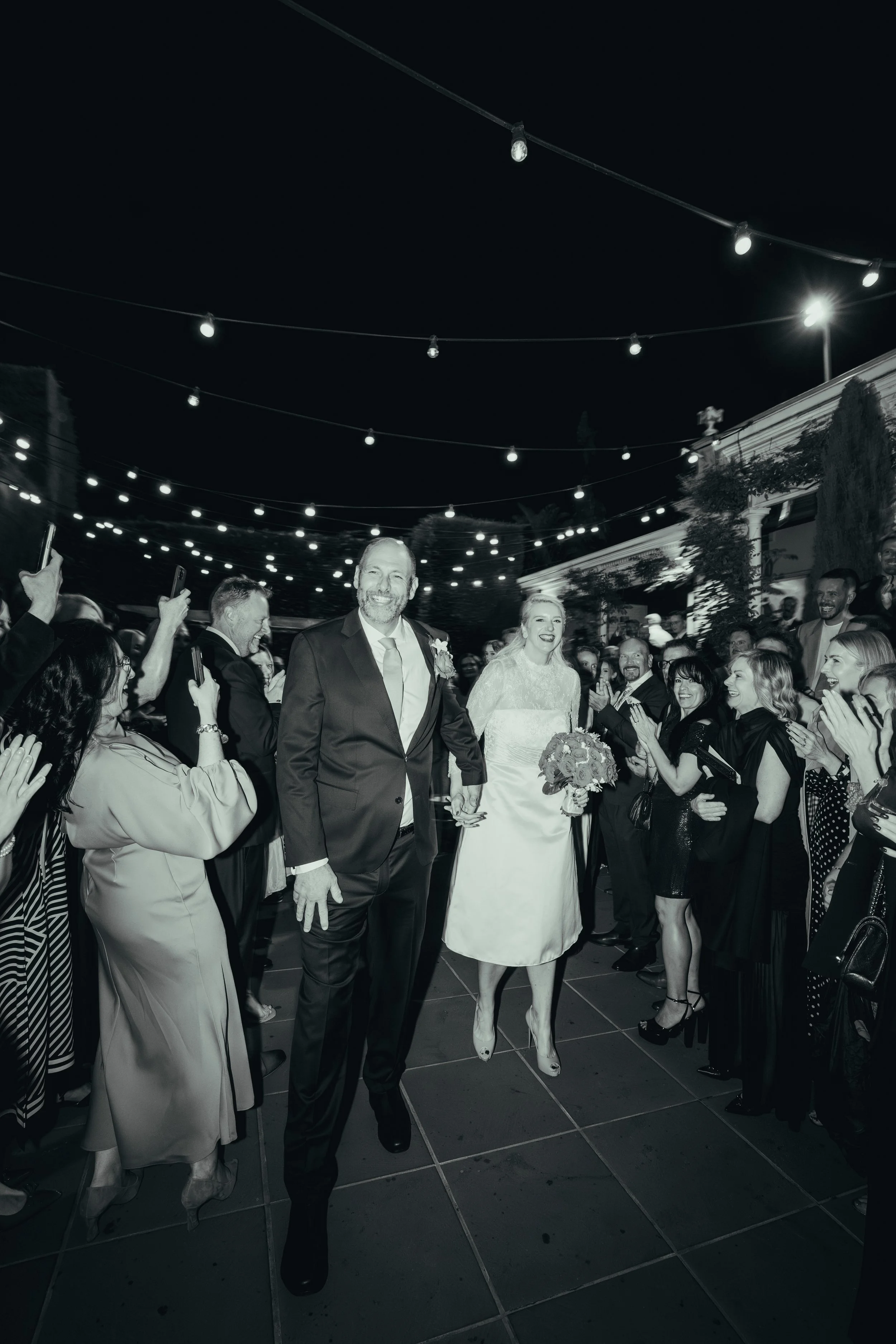 Wedding reception with bride and groom walking through a crowd outdoors at night under string lights, surrounded by happy guests.