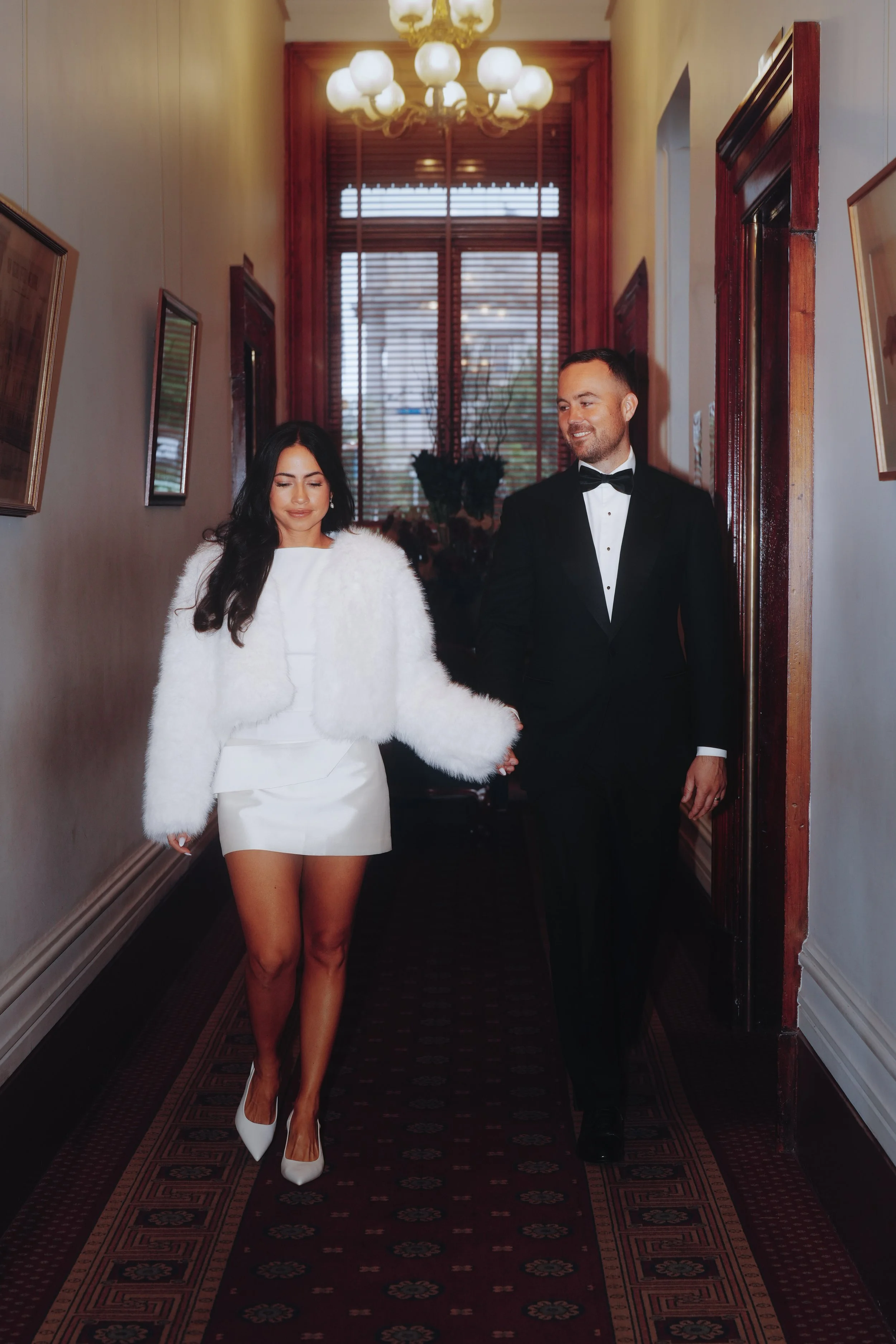 A woman in a white dress and fur coat holding hands with a man in a tuxedo walking down a hallway with framed pictures on the walls and a chandelier overhead.