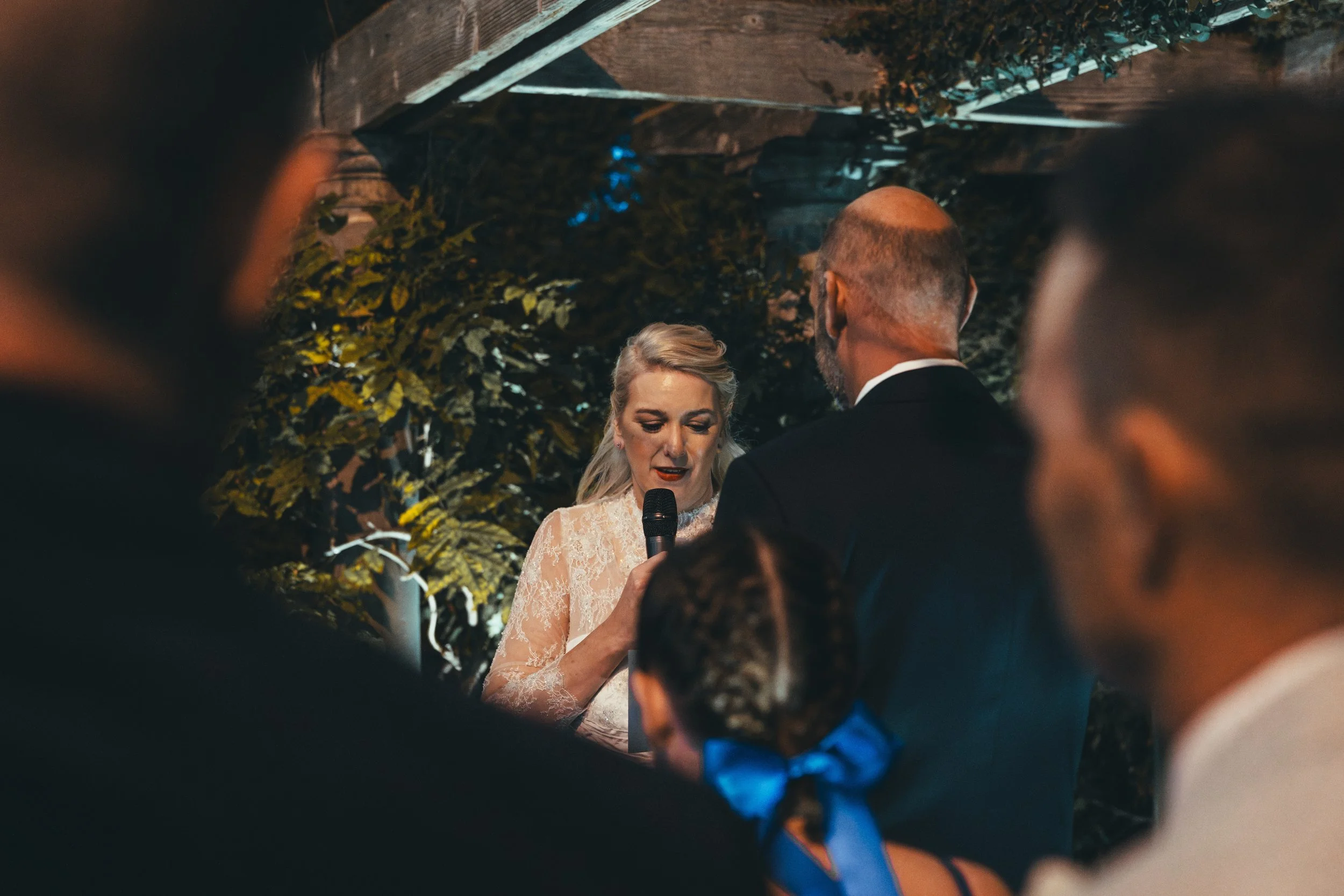 A woman in a white lace dress is reading vows during a wedding ceremony, held outdoors at night. She is holding a microphone, with a man in a dark suit standing in front of her. The scene is framed by blurred guests in the foreground and greenery in 