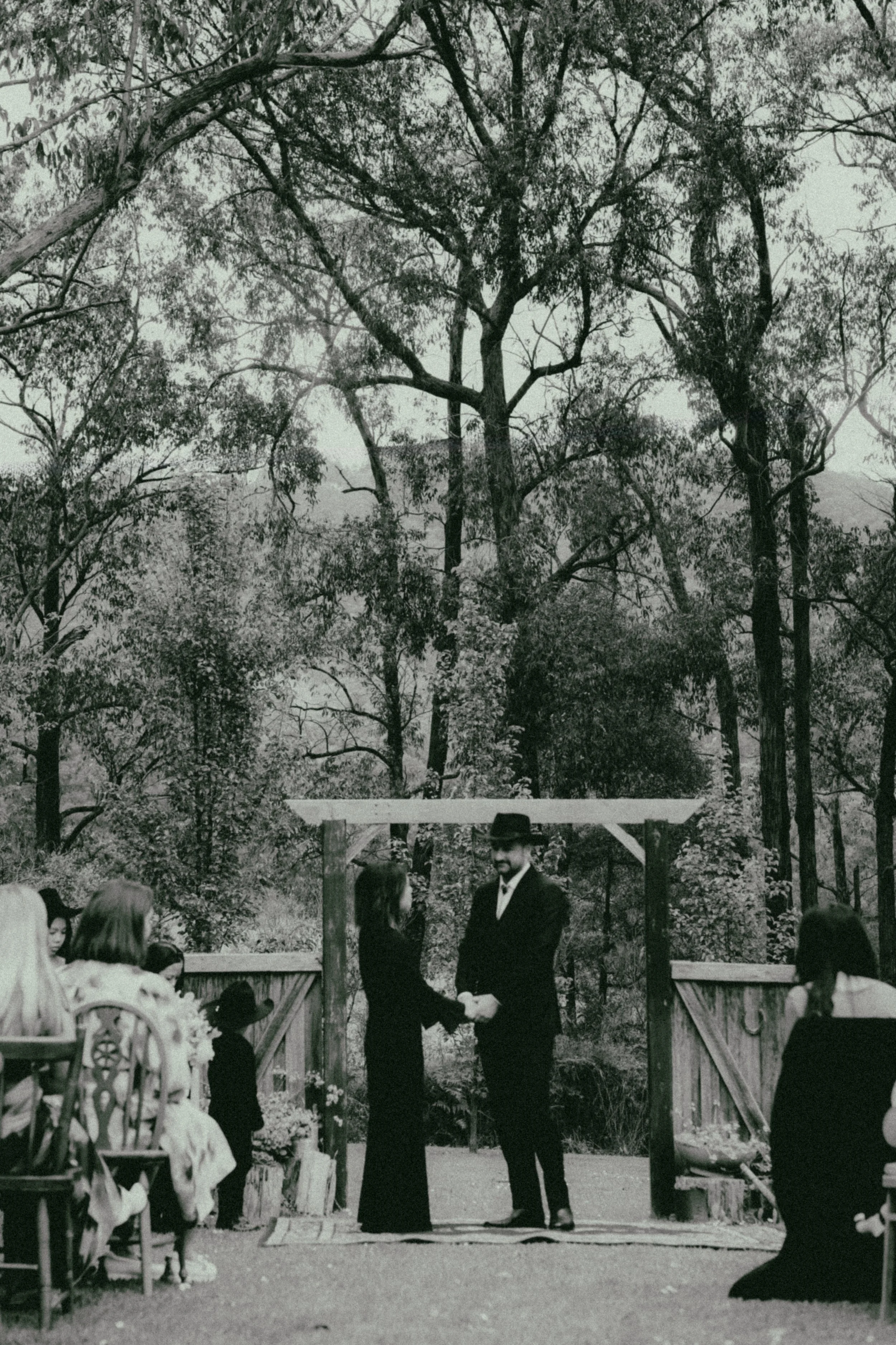 A wedding ceremony taking place outdoors in a wooded area. A bride and groom are exchanging vows in front of a small audience, with guests seated on either side. The background features tall trees and a rustic wooden arch.