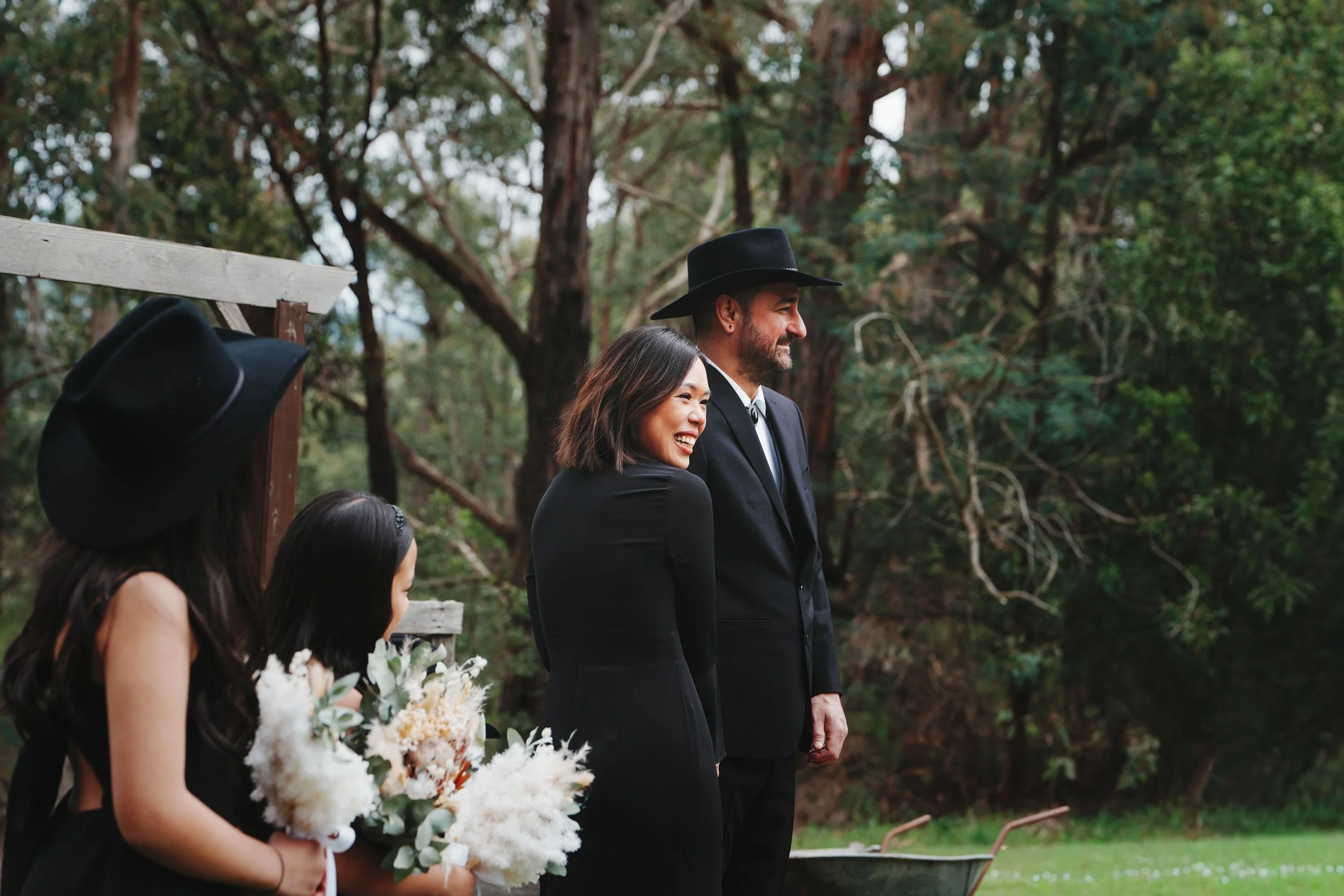 Group of four people outdoors, the woman with long dark hair holding a bouquet of white flowers, smiling, two men dressed in black suits and cowboy hats standing, surrounded by green trees.