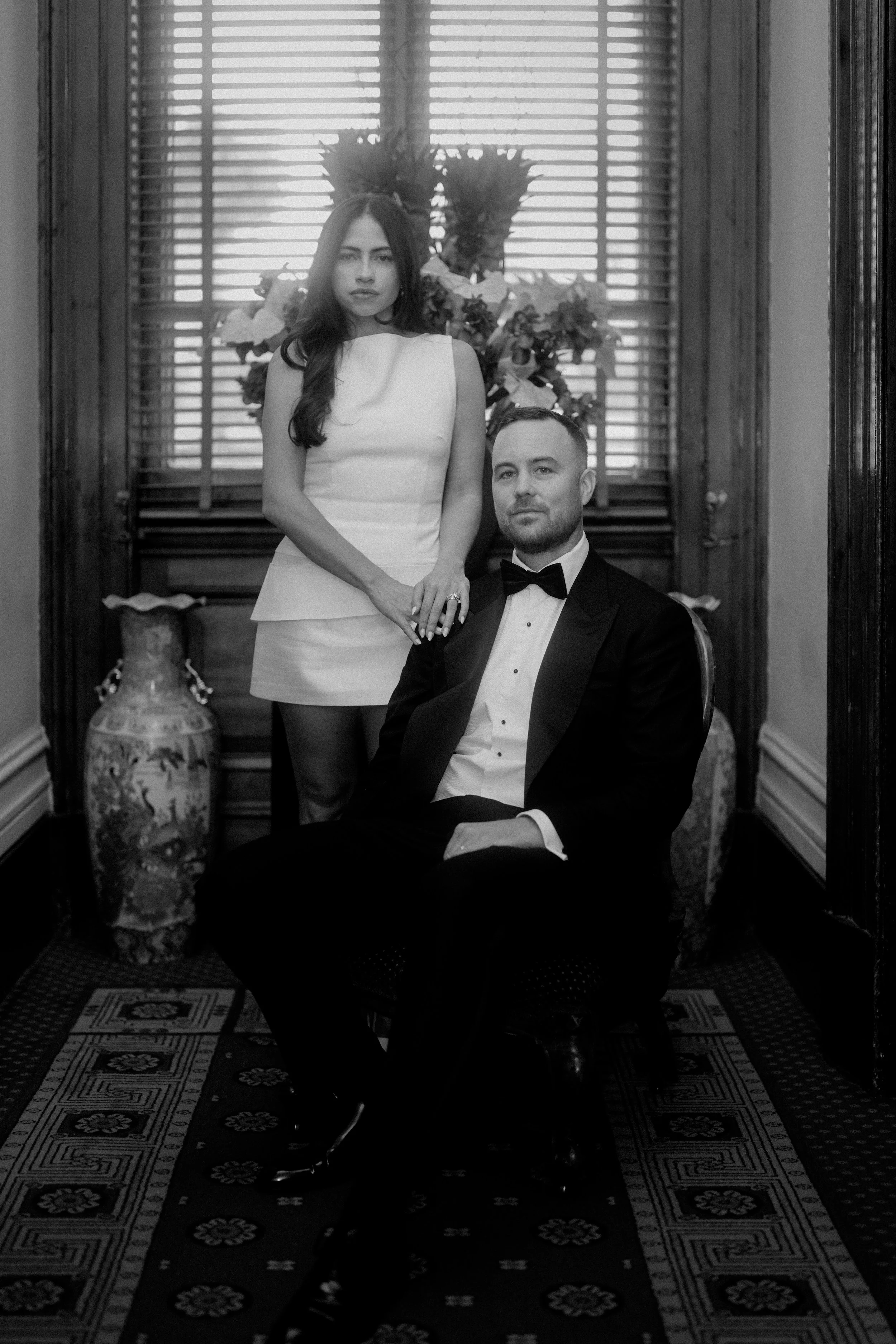 Black and white photo of a woman in a white dress standing behind a seated man in a tuxedo, in a room with wooden window blinds and floral decorations.
