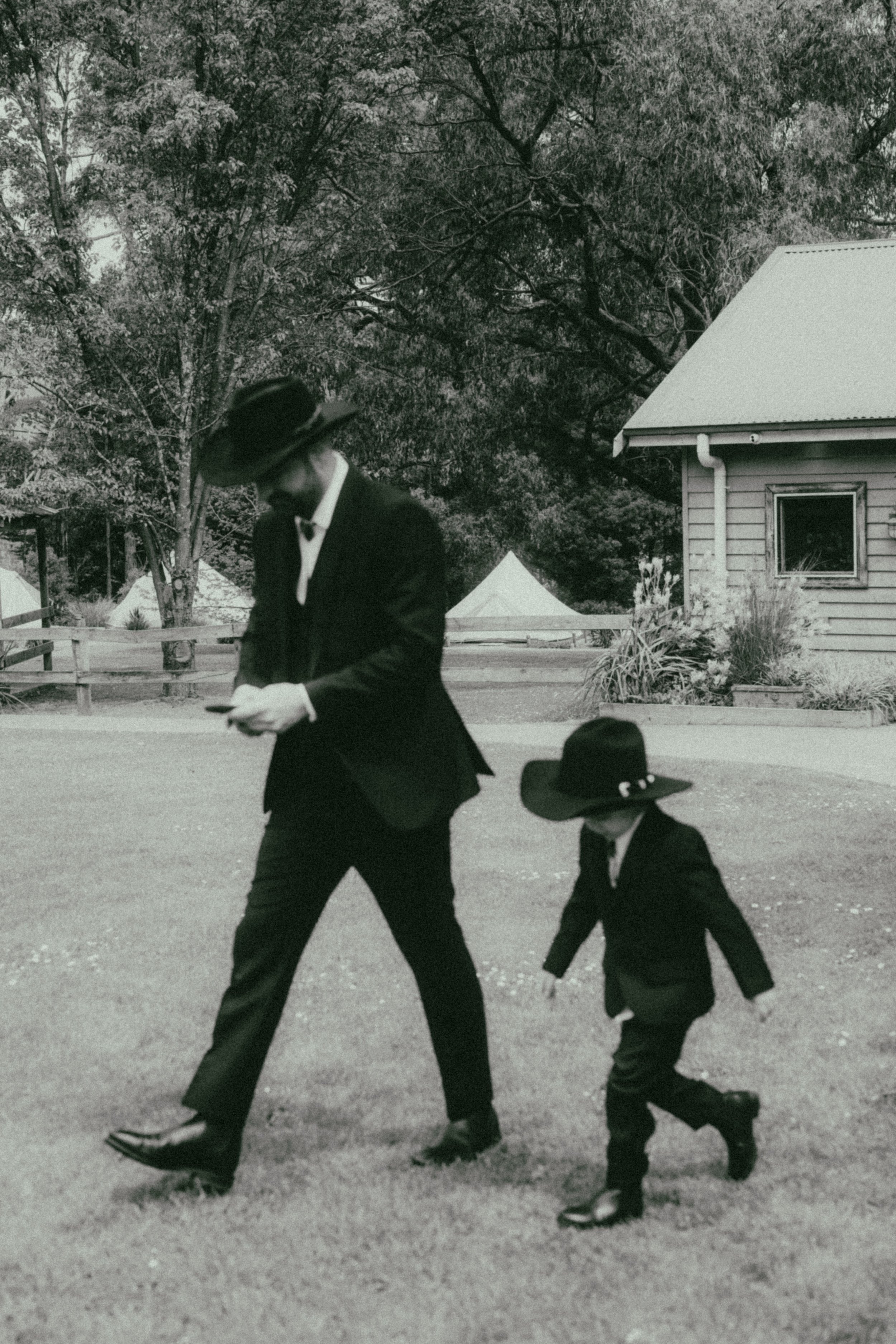 A black-and-white photo of a man and a young boy walking, dressed in dark suits, hats, and shoes. They are outdoors near a house with trees and a fence in the background.