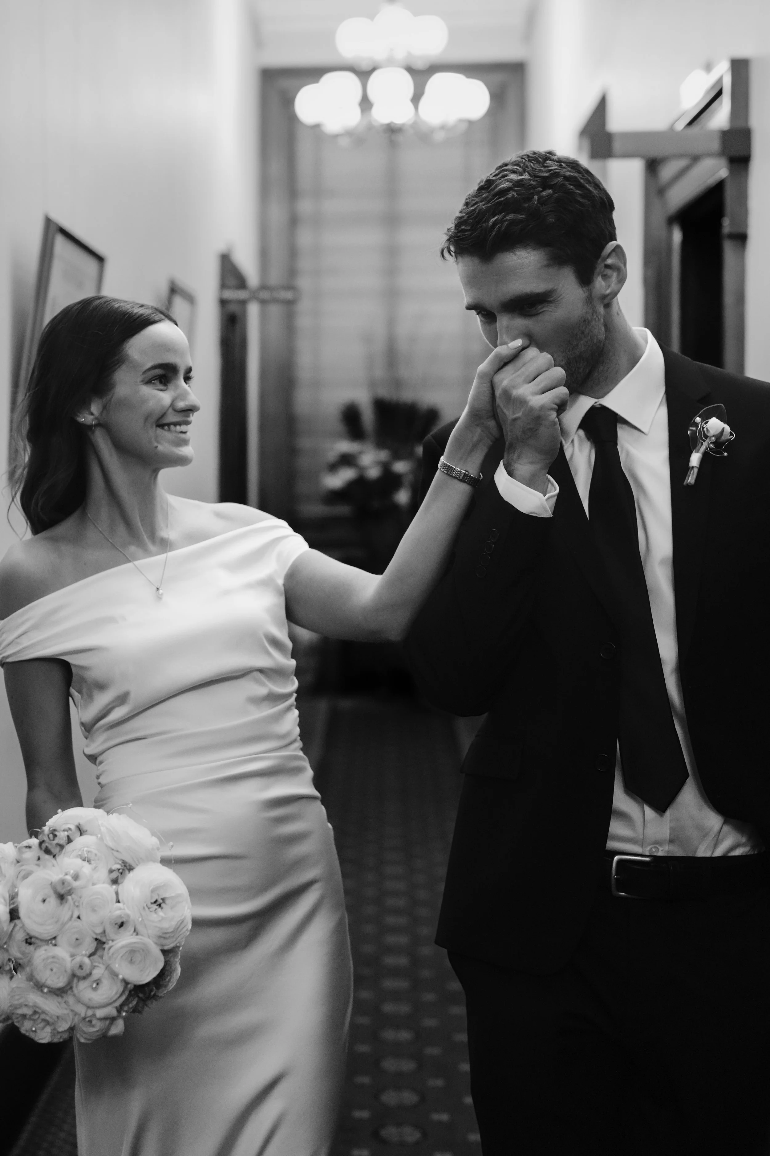 A black-and-white photo of a bride and groom at their wedding. The bride, smiling, is holding a bouquet of roses and wearing a satin wedding dress. The groom, dressed in a dark suit with a white shirt and tie, is smiling and touching his nose with hi